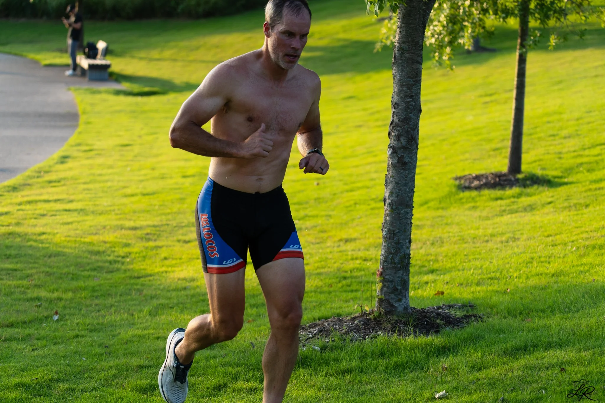 A shirtless man running outdoors in a park on a sunny day, wearing black and blue shorts and running shoes, with trees and a grassy area in the background.