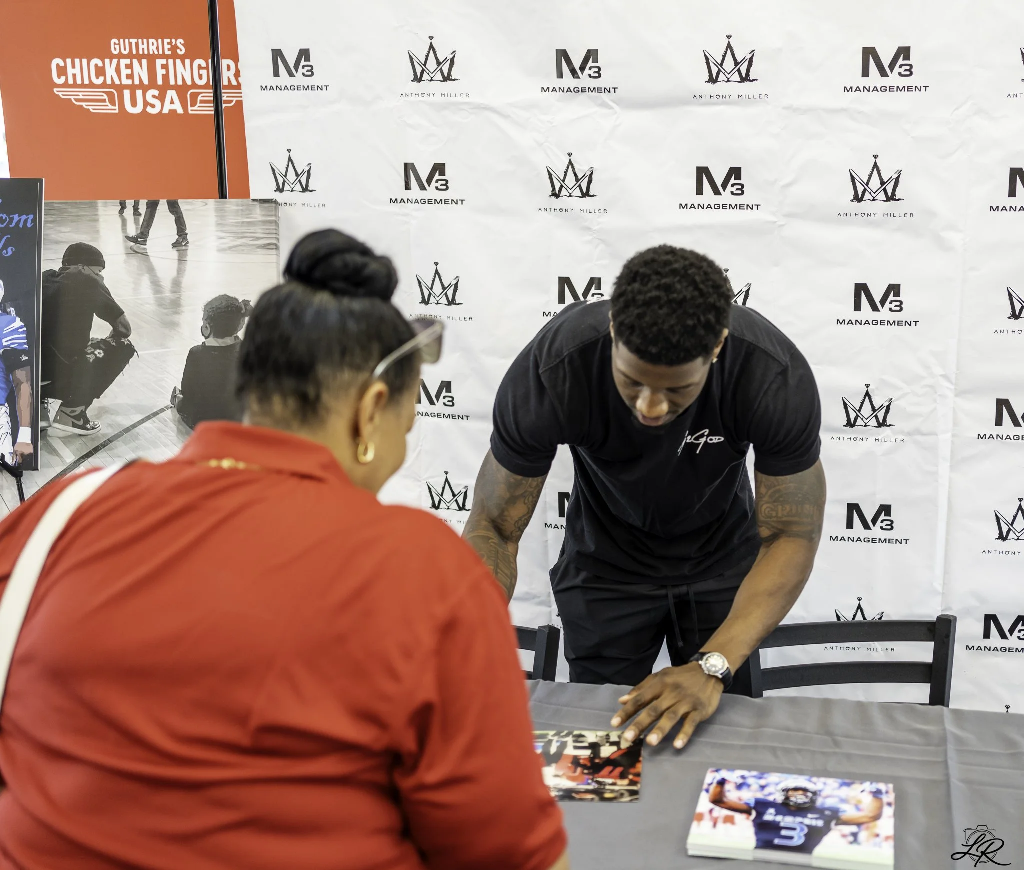 People at a signing event with a backdrop displaying logos of M3 Management, Anthony Miller, and a crown symbol. A man in black is signing items at a table while others watch.
