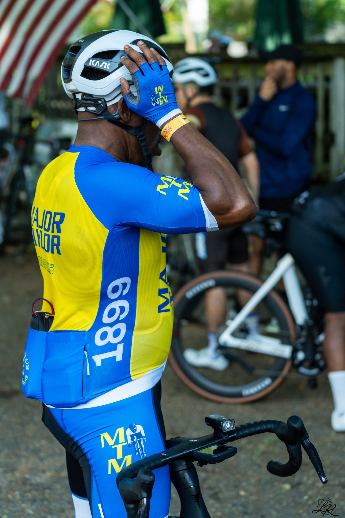 A cyclist in a blue and yellow jersey holding a white bike helmet to his head, with a group of people and a bicycle in the background.