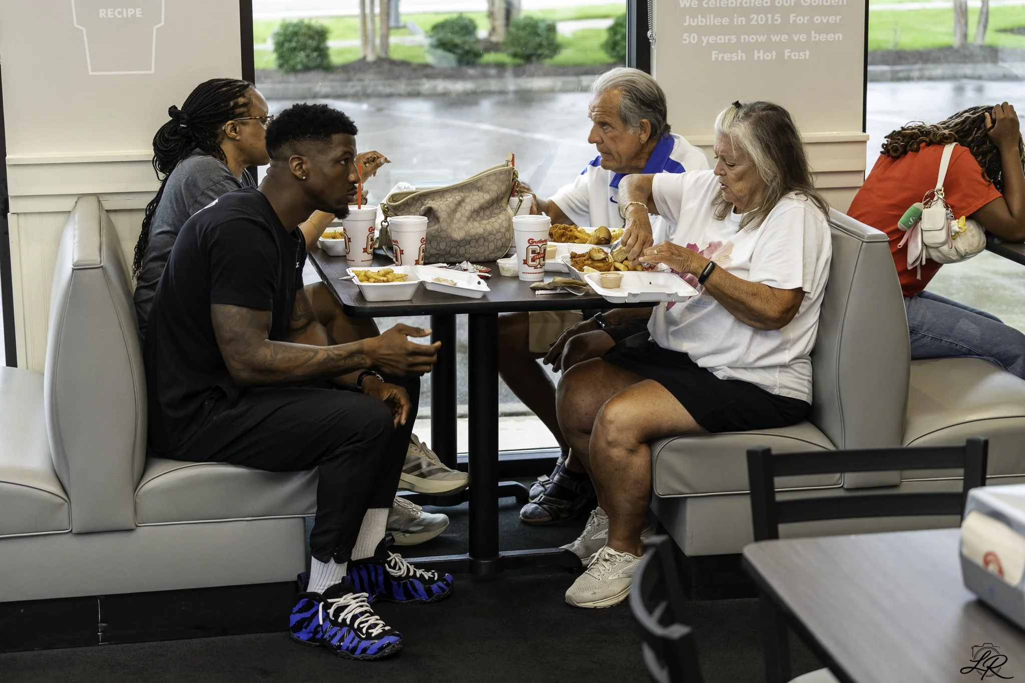 Five people sit at a table inside a restaurant enjoying a meal. Two older women and three younger people, including a man with tattoos and a woman with braided hair. They have fast food plates, drinks, and a purse on the table. The restaurant has lar