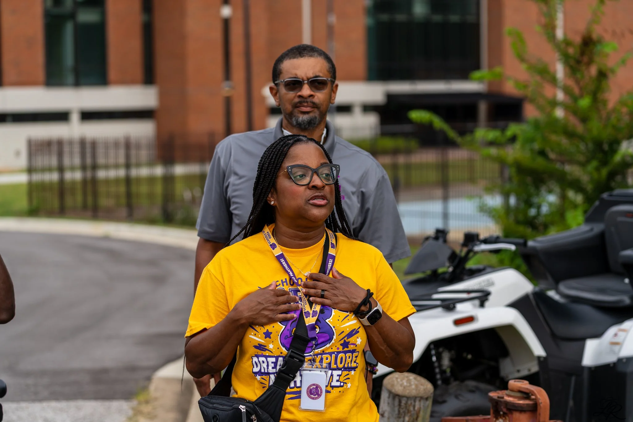 A woman with glasses, wearing a yellow T-shirt, stands with her hands on her chest, surrounded by two lanyards and a camera, during an outdoor event, with a man standing behind her in front of a building and a golf cart nearby.