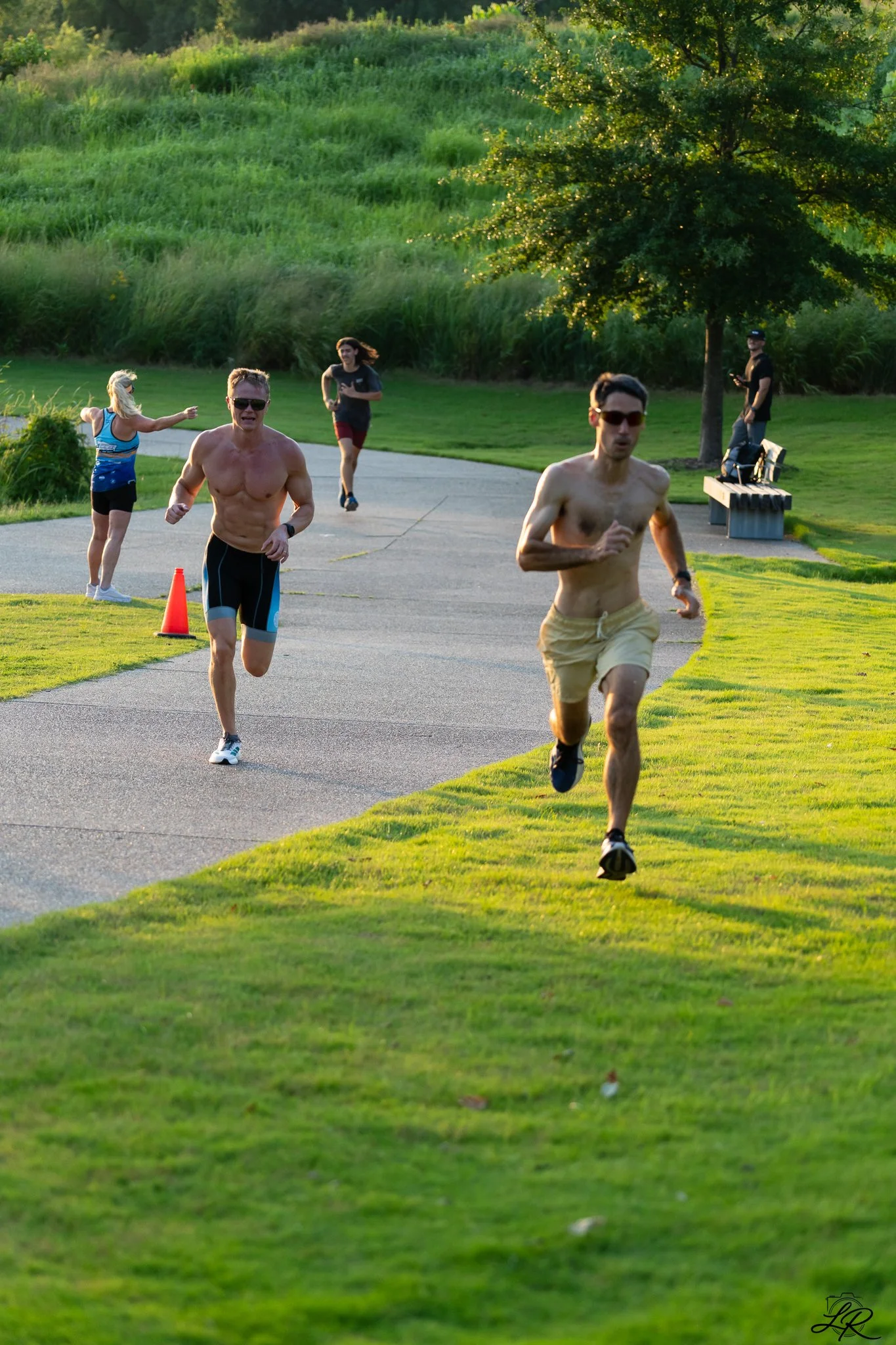 Group of people running outdoors on a paved pathway in a park during sunset, with lush green grass and trees in the background.