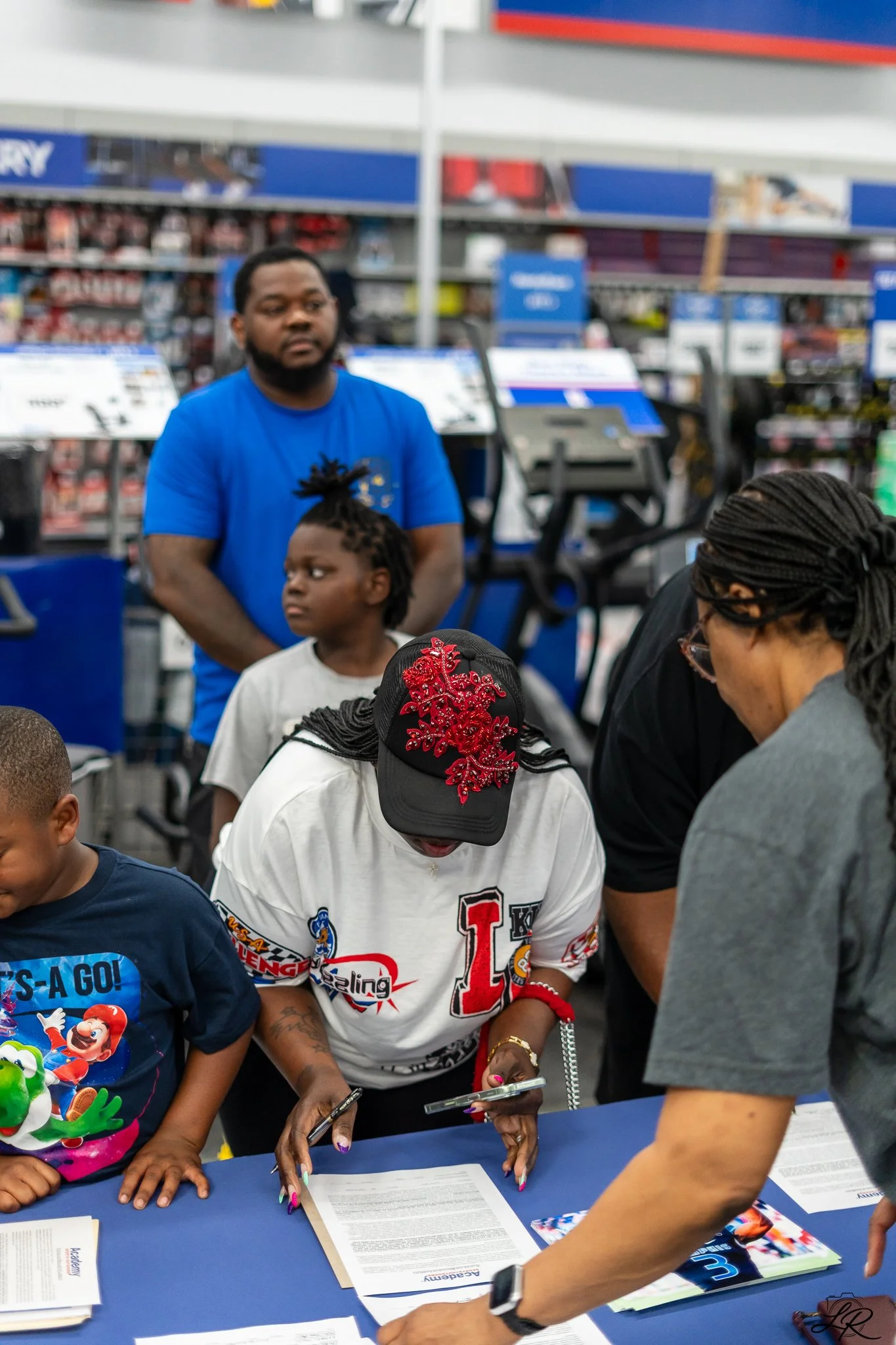 A group of people gathered around a table at a sign-up or registration event inside a store, with shelves and products visible in the background.