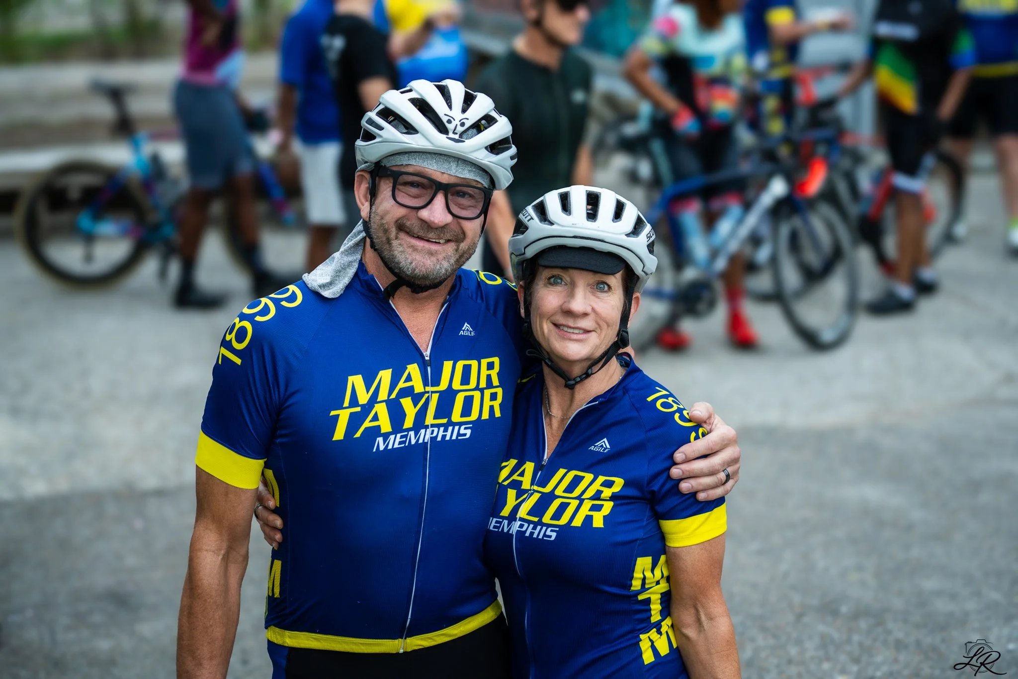 A man and woman wearing blue athletic jerseys with yellow accents and the words "Major Taylor Memphis" on the front, smiling for the camera while standing arm-in-arm outdoors, with a group of cyclists and blurred trees in the background.