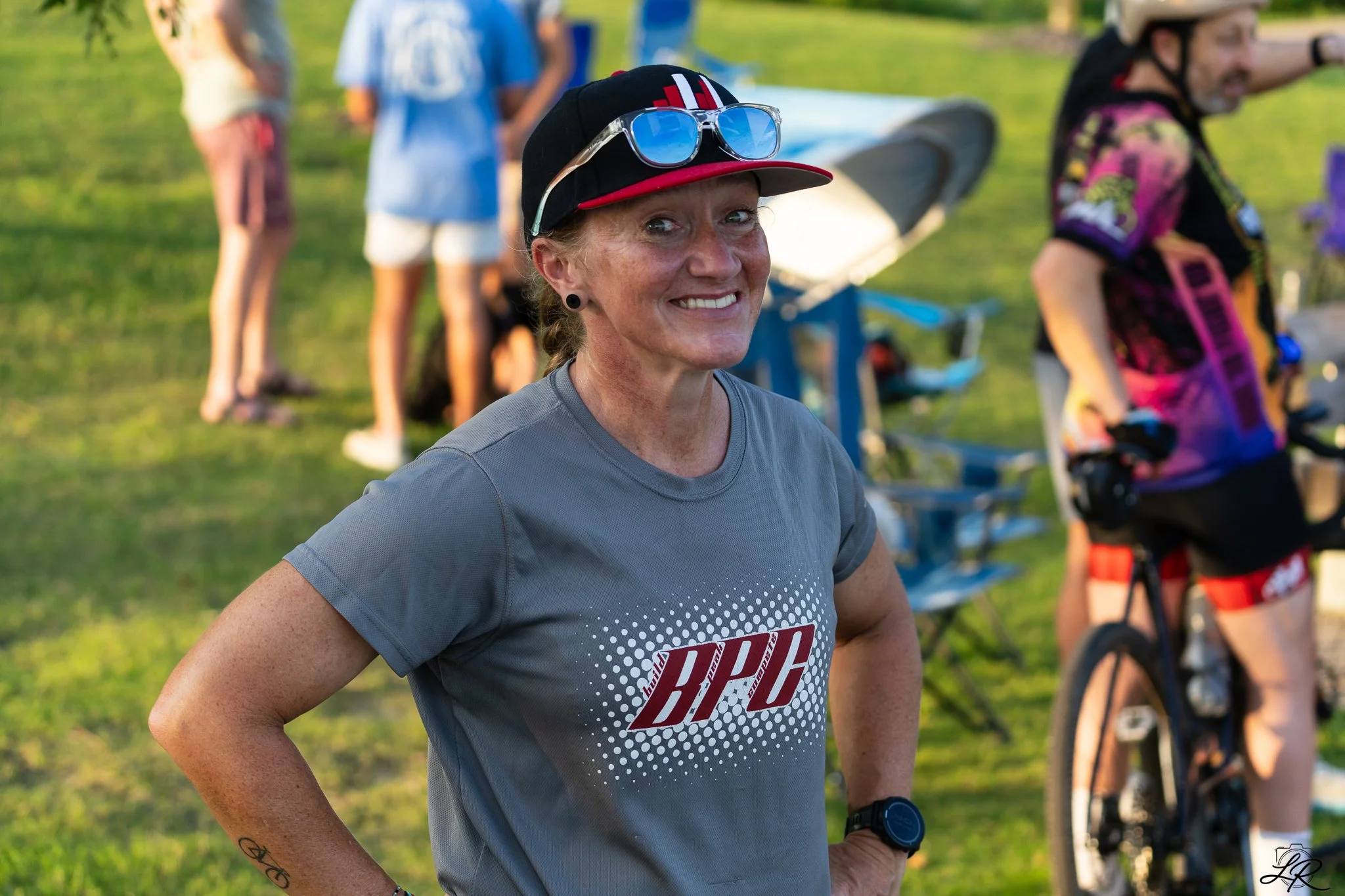 A smiling woman with light skin, freckles, and blonde hair, wearing a black cap with sunglasses on it, a gray athletic shirt with red and white text, and a smartwatch, standing outdoors at a park with other people and bicycles in the background.