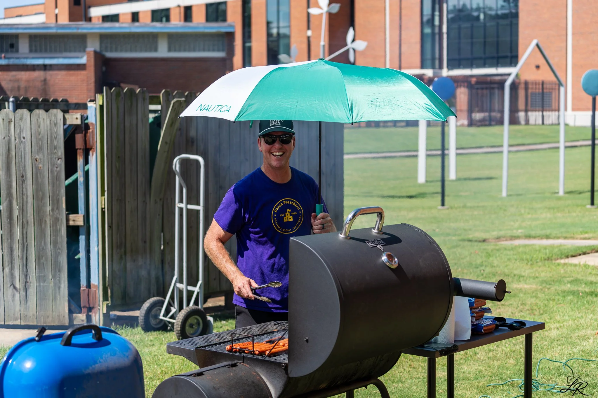 A man in sunglasses and a navy T-shirt is standing next to a black charcoal grill, cooking hot dogs and sausages. He is holding a pair of tongs and smiling, with a green and white Nautica branded umbrella overhead. Outside on a grassy area, there is 