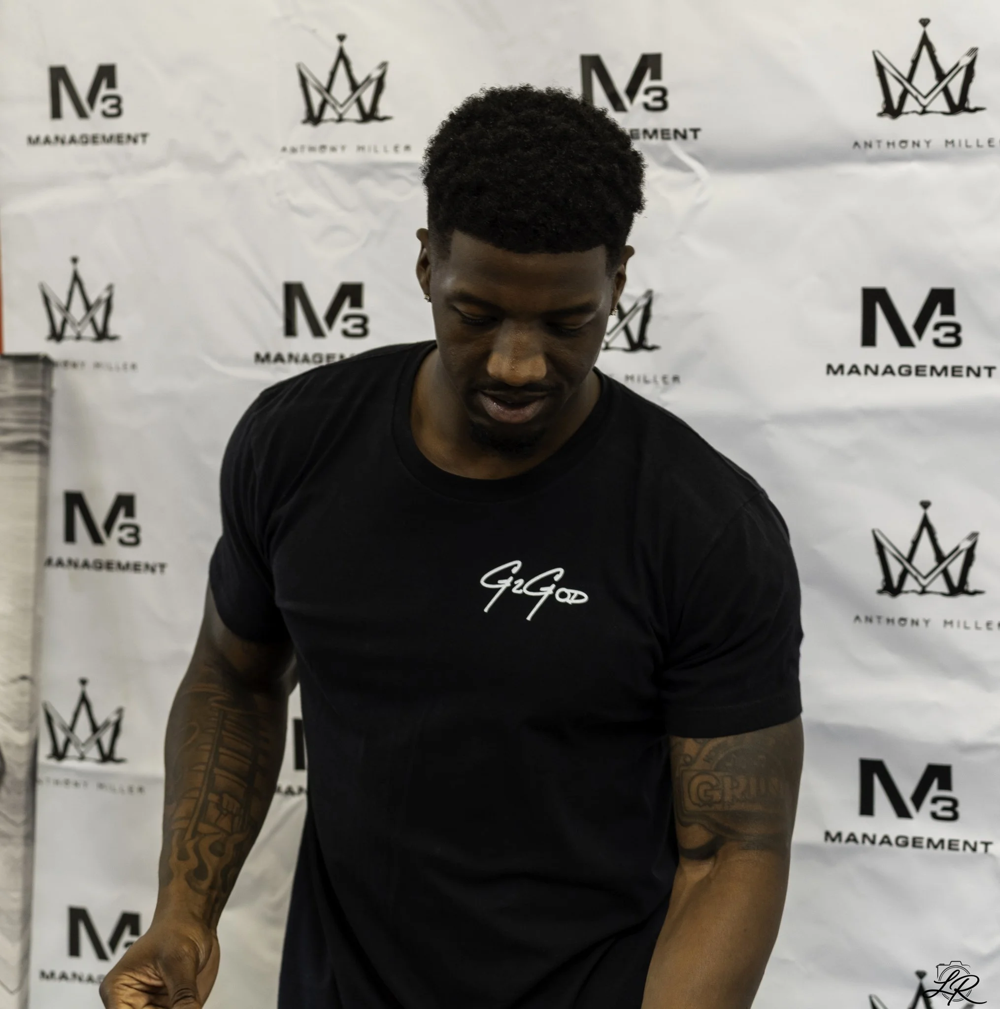 A person with tattoos on their arms looking down in front of a step and repeat backdrop with the text 'Management' and the name 'Anthony Miller' and a crown logo.
