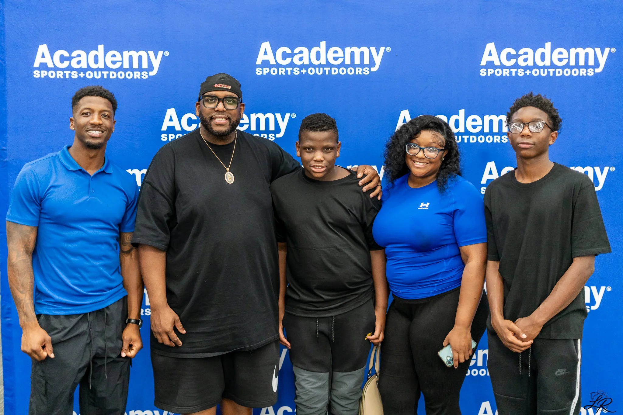 Group of five individuals standing together with their arms around each other, smiling, in front of a blue backdrop with white 'Academy Sports + Outdoors' logos.