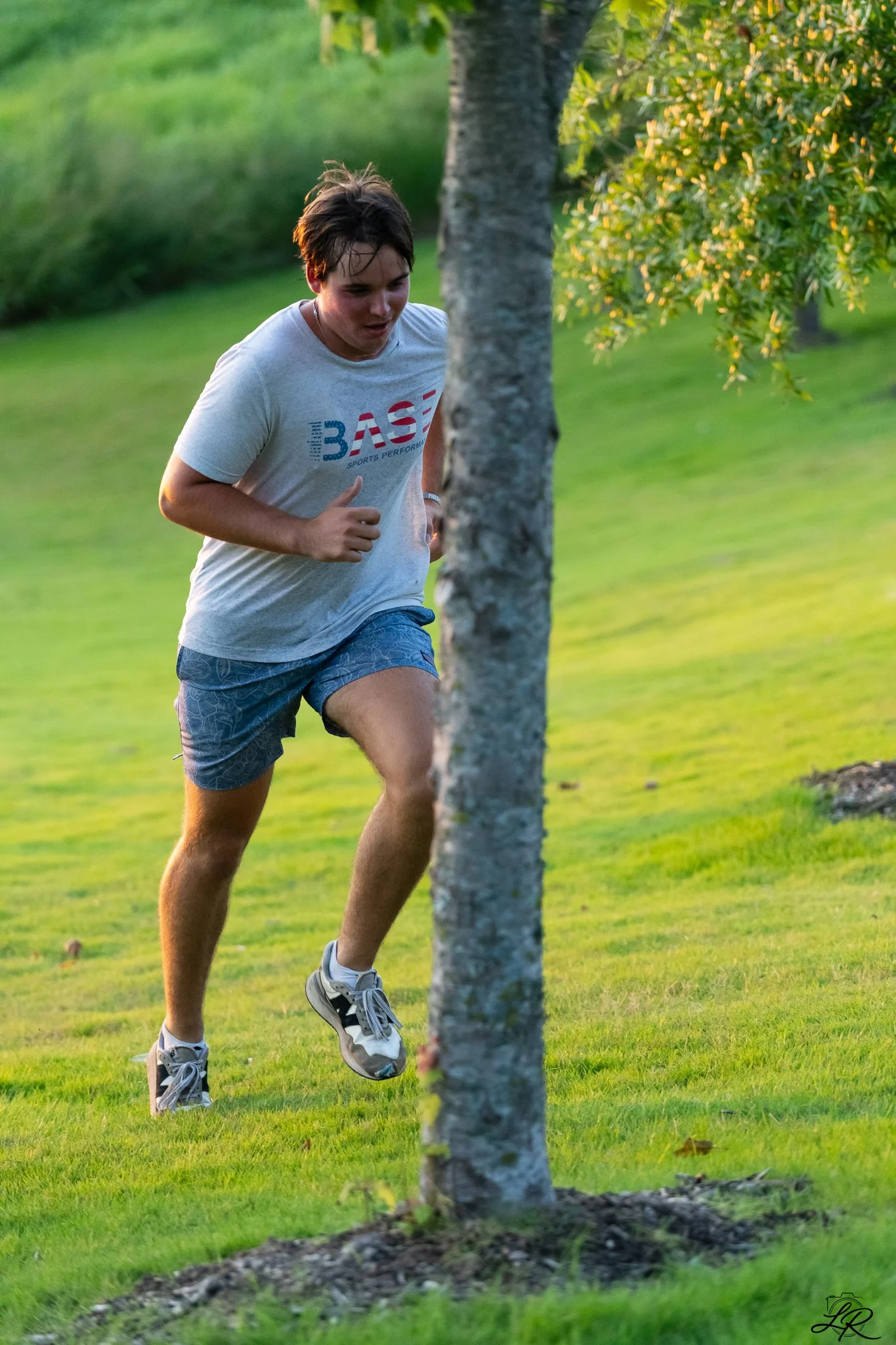 A teenage boy running outdoors on a grassy field with trees, wearing a white t-shirt and blue shorts.