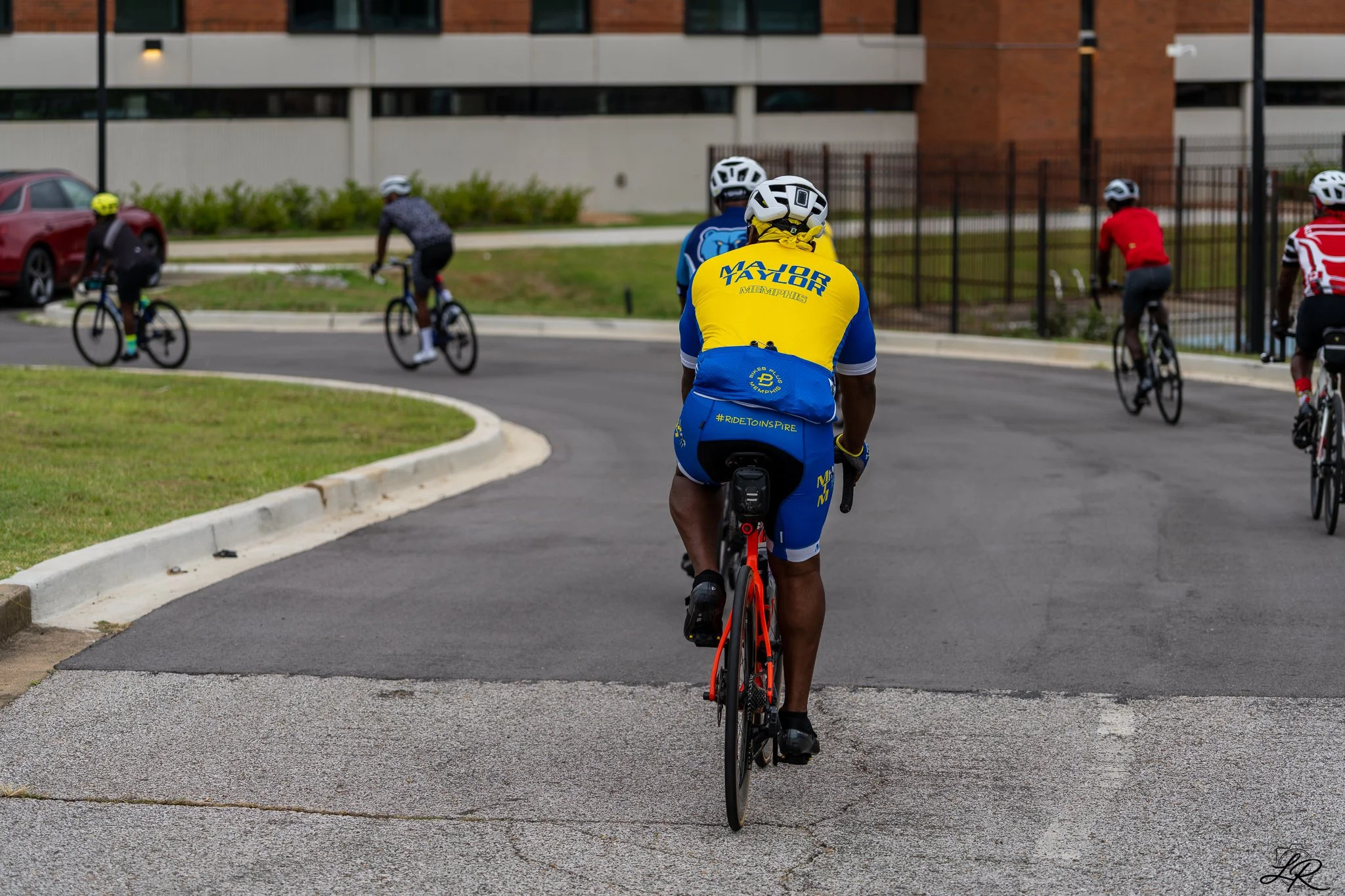 A group of people riding bicycles on a paved path in an urban area, with a building, a fence, and parked cars in the background.