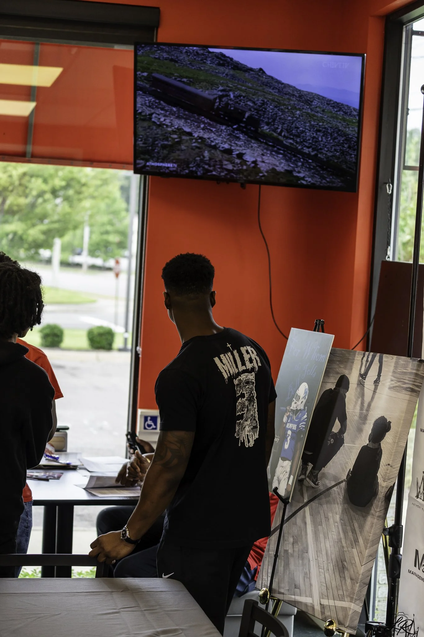 People attending a registration or check-in table at an event, with posters, informational displays, and a TV screen showing a nature scene, in a room with large windows and red walls.