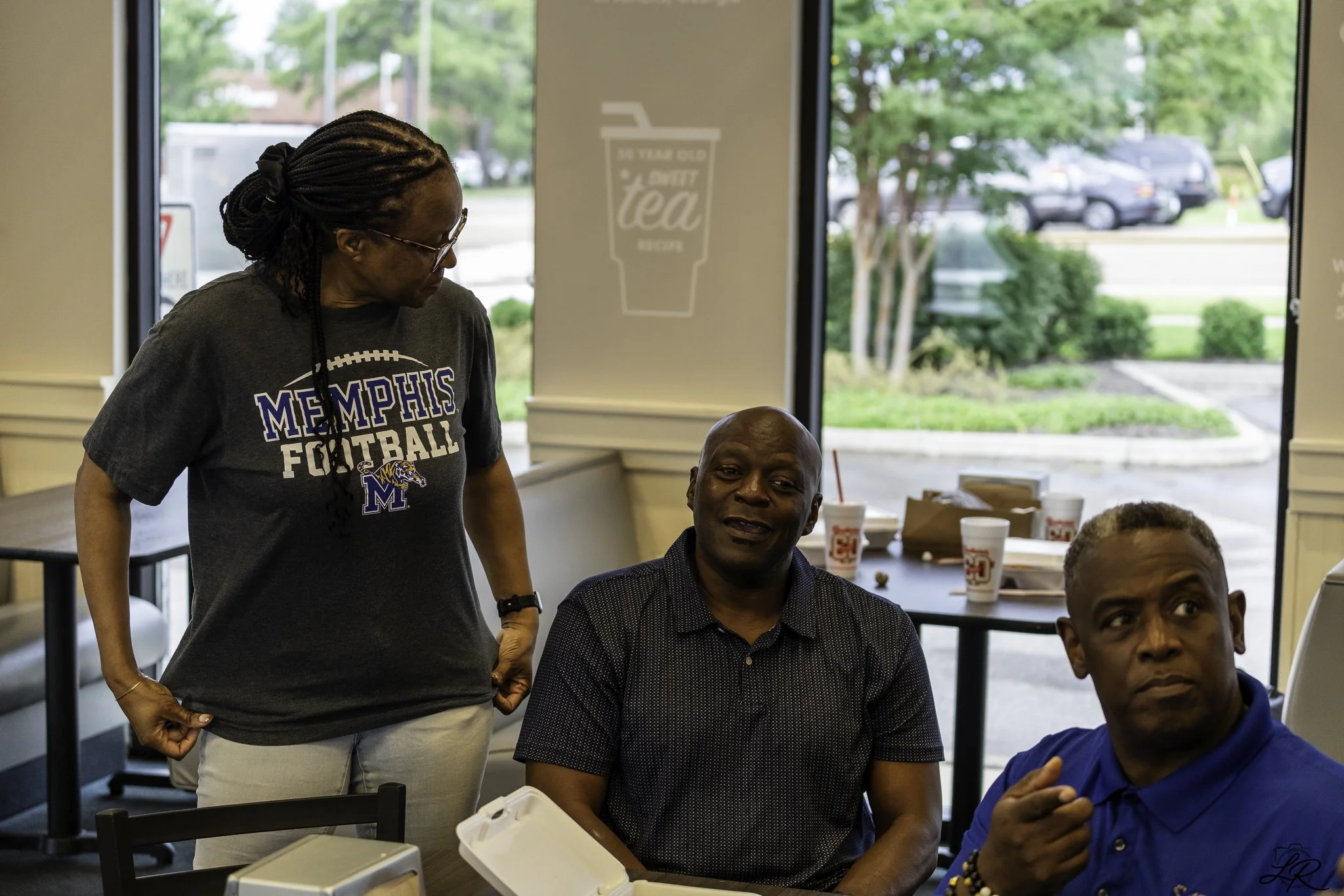People gathered around a table inside a cafe or restaurant, with large windows showing trees and parked cars outside. One woman with glasses and a Memphis Football shirt stands beside a seated man, while other individuals are partially visible around