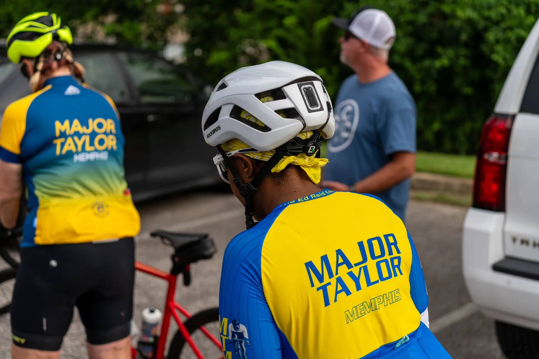 Group of cyclists wearing yellow and blue jerseys, with one helmet in the foreground, at a biking event with a support team in the background.