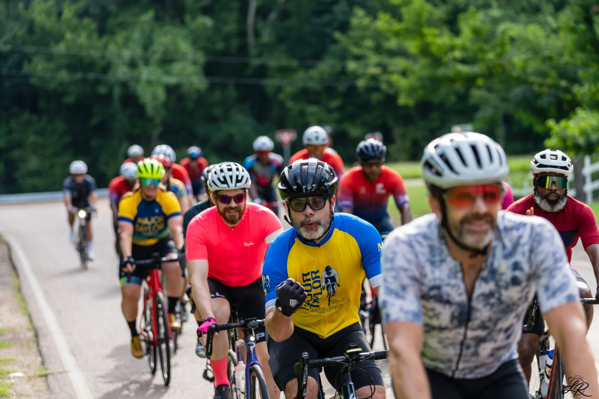 A group of diverse cyclists wearing helmets and athletic clothing riding on a paved road through a green, wooded area.