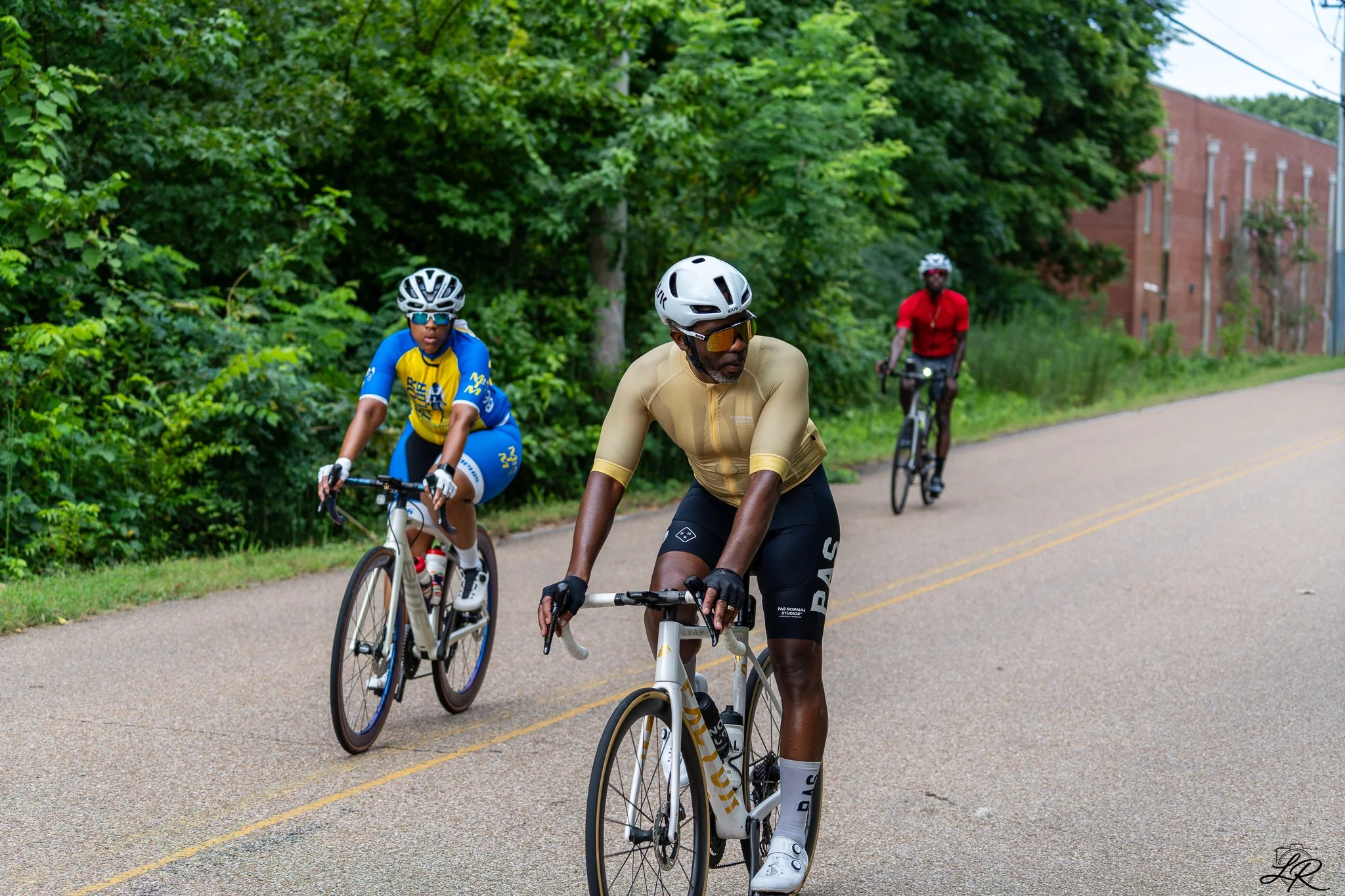 Three cyclists riding on a paved road lined with green trees and a brick building in the background. The cyclist in front is wearing a tan cycling outfit and sunglasses, the middle cyclist is wearing a blue and yellow jersey with a white helmet and s