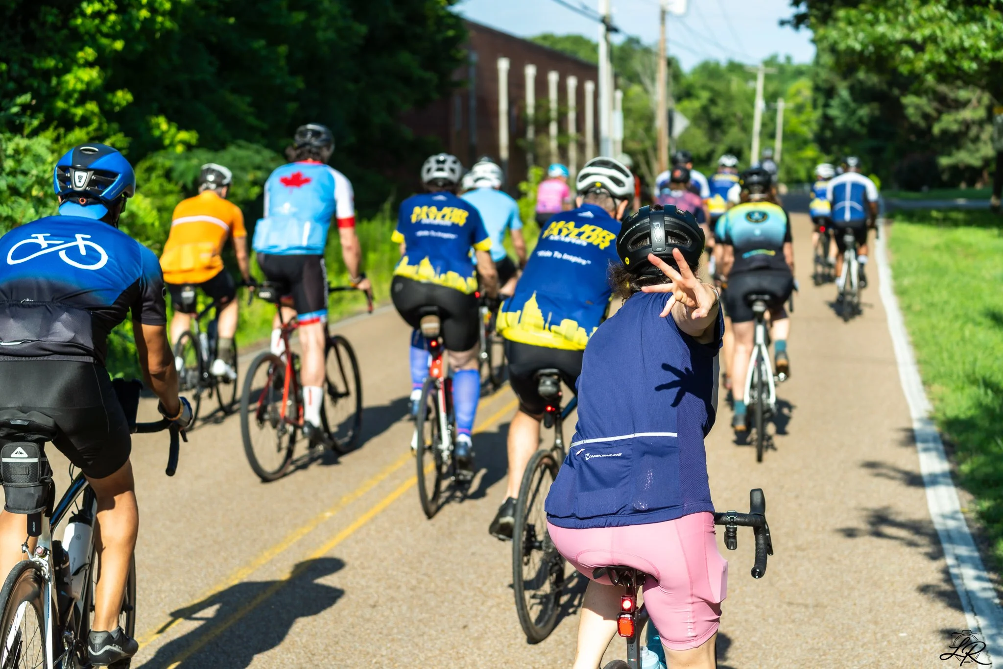 A group of people riding bicycles on a rural road with greenery on the sides, wearing helmets and colorful athletic clothing. One person in the foreground is making a peace sign.
