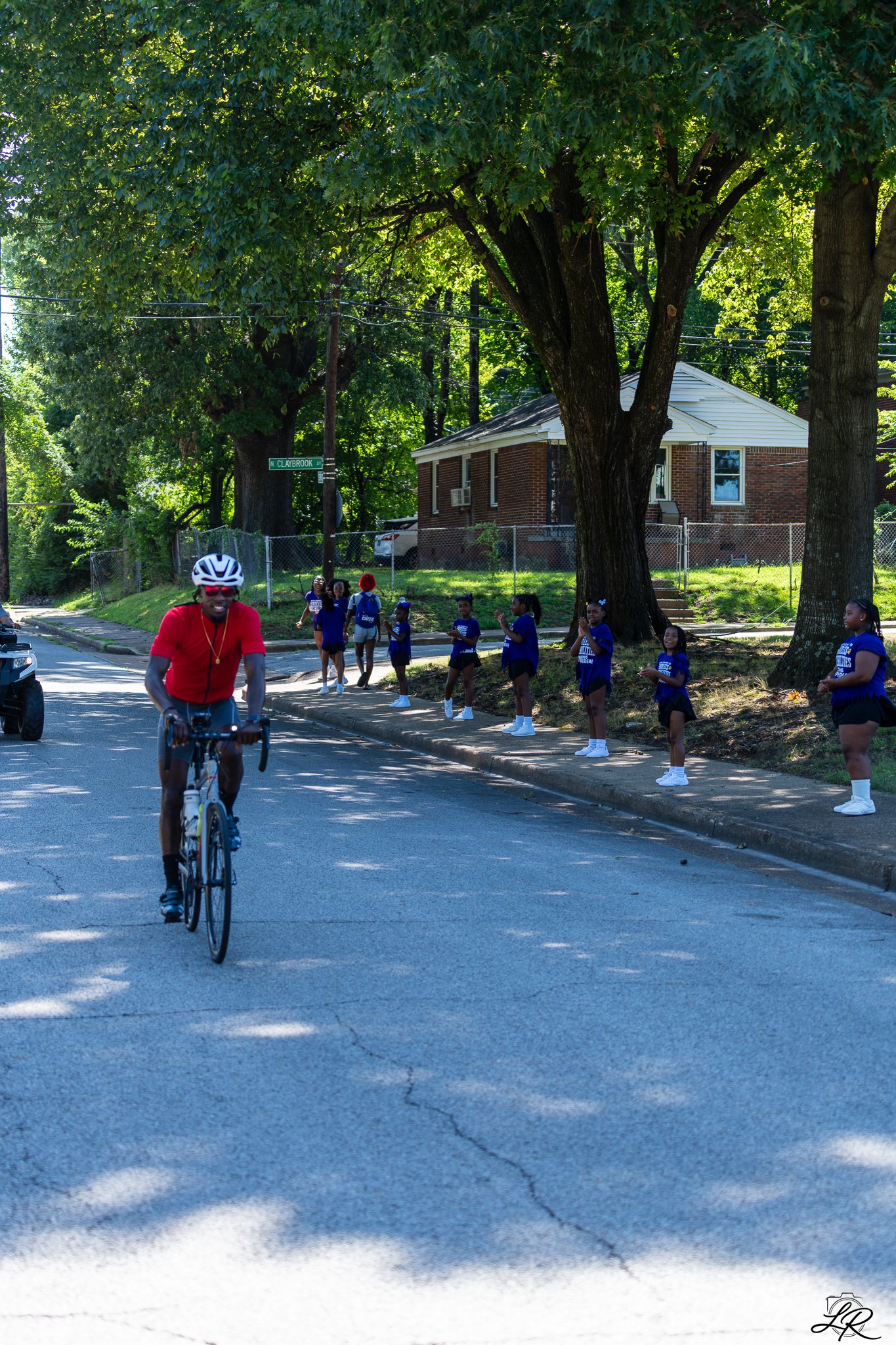 A man in a red shirt and helmet riding a bicycle on a street with children standing by the sidewalk, some clapping. Large trees and a house are in the background. The street sign reads 'N Claybrook St'.