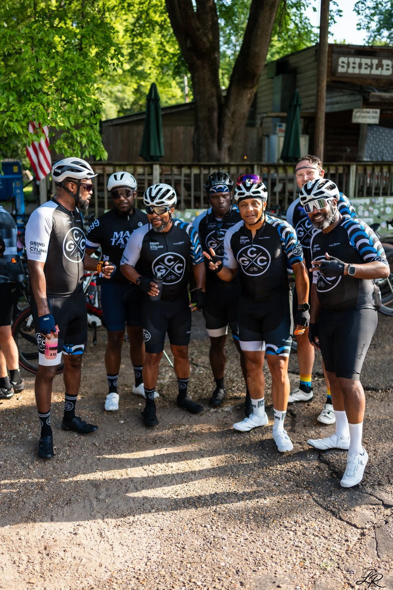 A group of six men in cycling gear, wearing helmets and sunglasses, standing together outdoors in front of a wooden fence and a building, with trees and an American flag in the background.