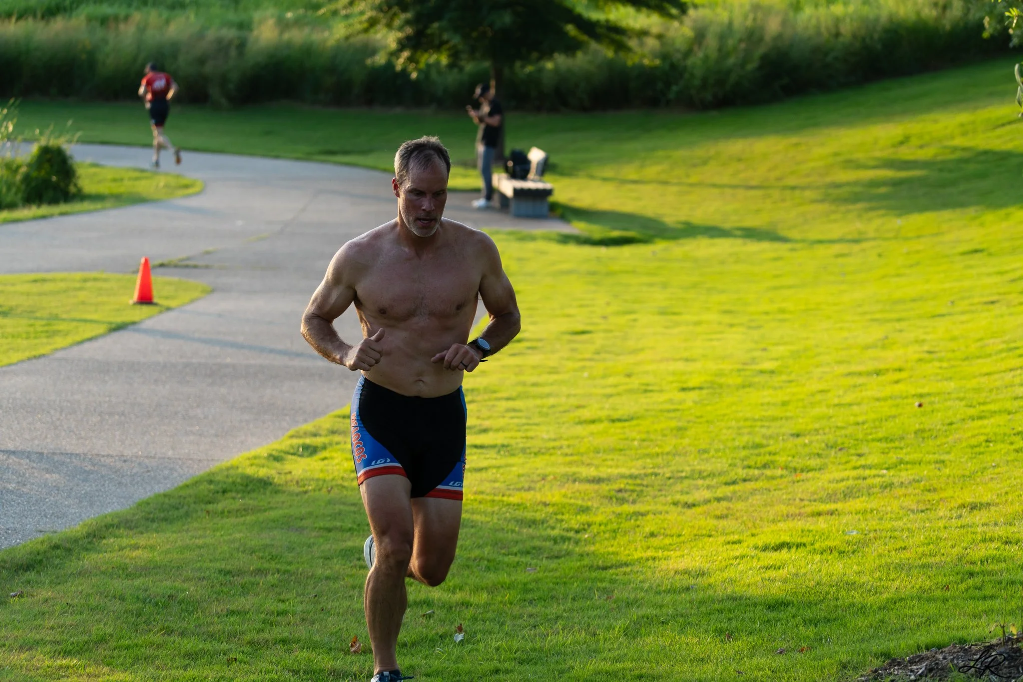 A man running outdoors on a grassy path, shirtless, wearing shorts and a watch, with trees and a person using a mobile phone in the background.