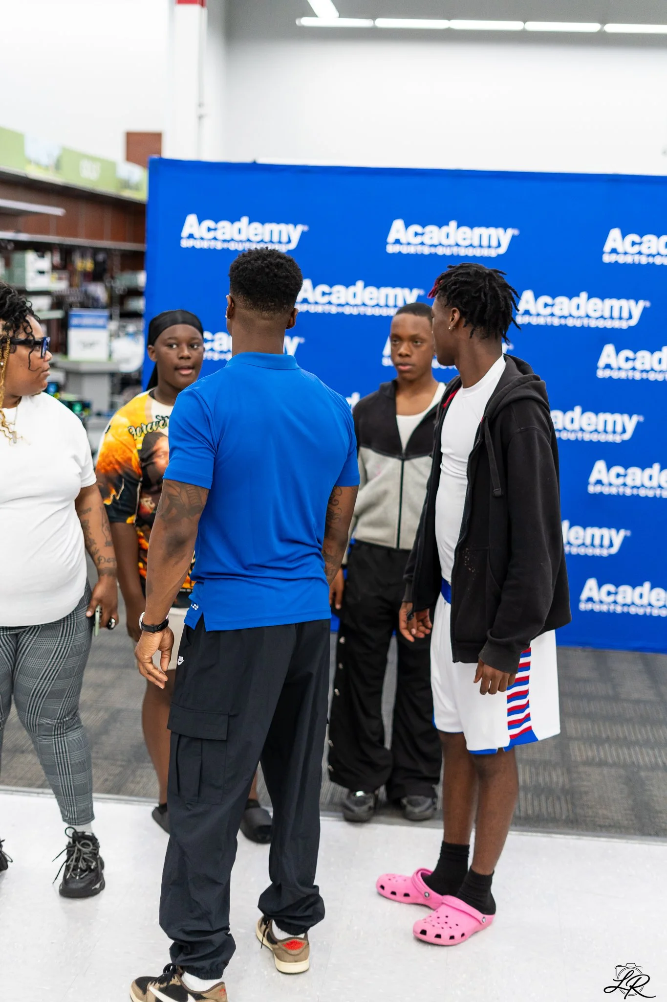 A group of five young people talking in front of a blue backdrop with the 'Academy Sports + Outdoors' logo. They are indoors, possibly at a store or event.