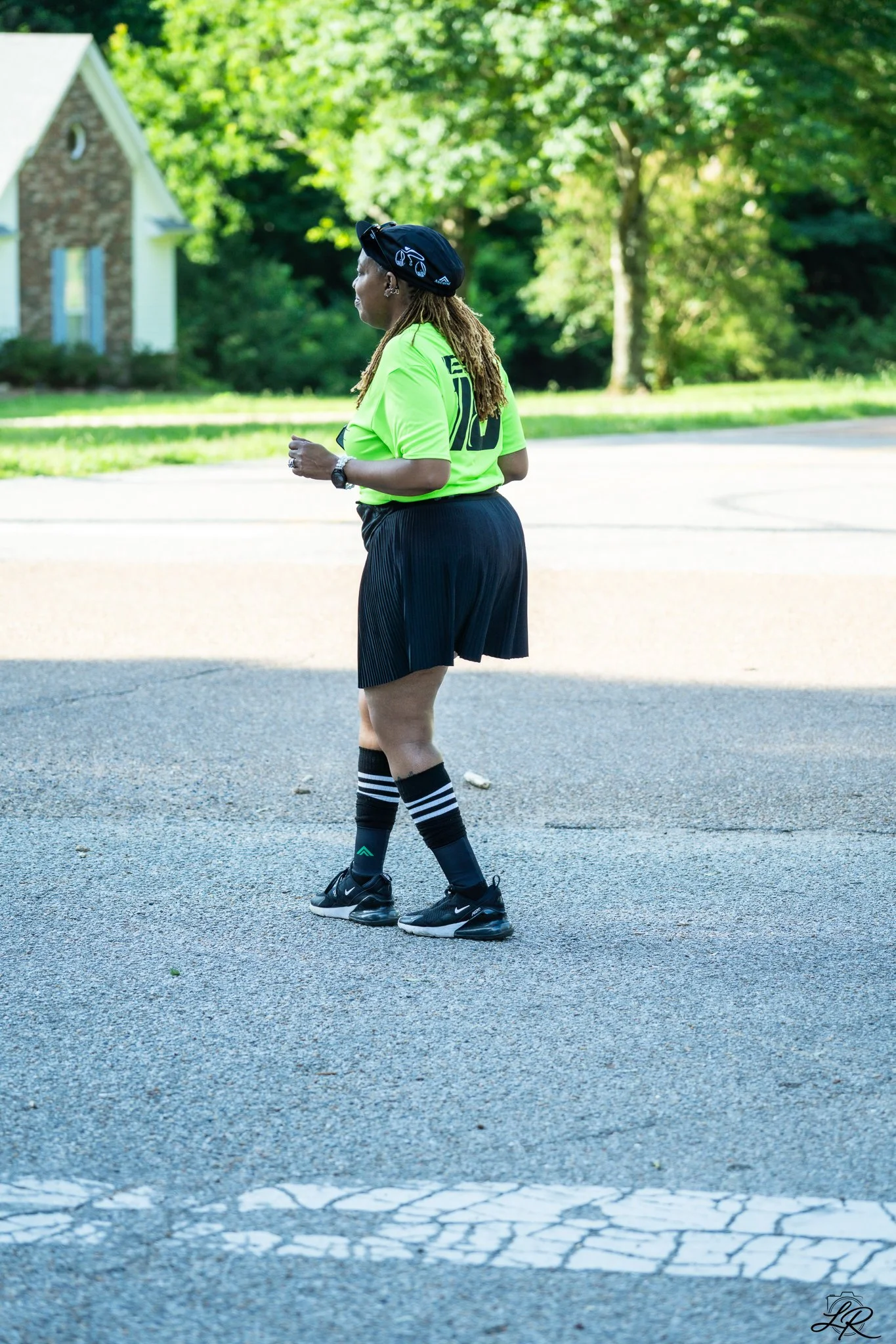 A woman with long dreadlocks wearing a neon green sports shirt, black shorts, black and white striped knee-high socks, black sneakers, a black cap, and a watch, is jogging on a paved street with greenery and houses in the background.