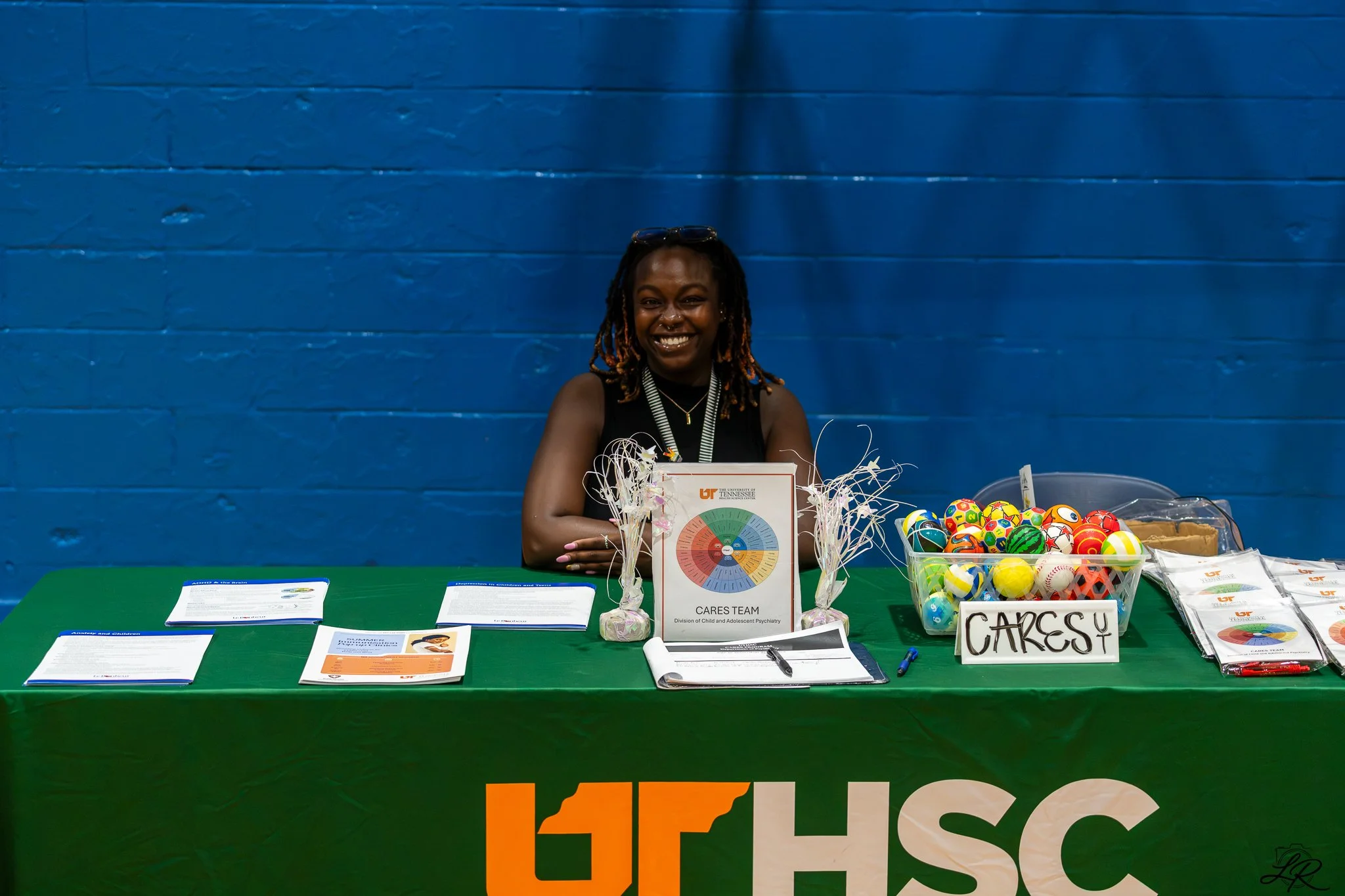 A woman with glasses on her head sitting behind a table at an event booth, smiling at the camera. The table displays informational pamphlets, a pie chart, decorative containers with wrapped candies, and a sign that reads 'CARES.' The background featu