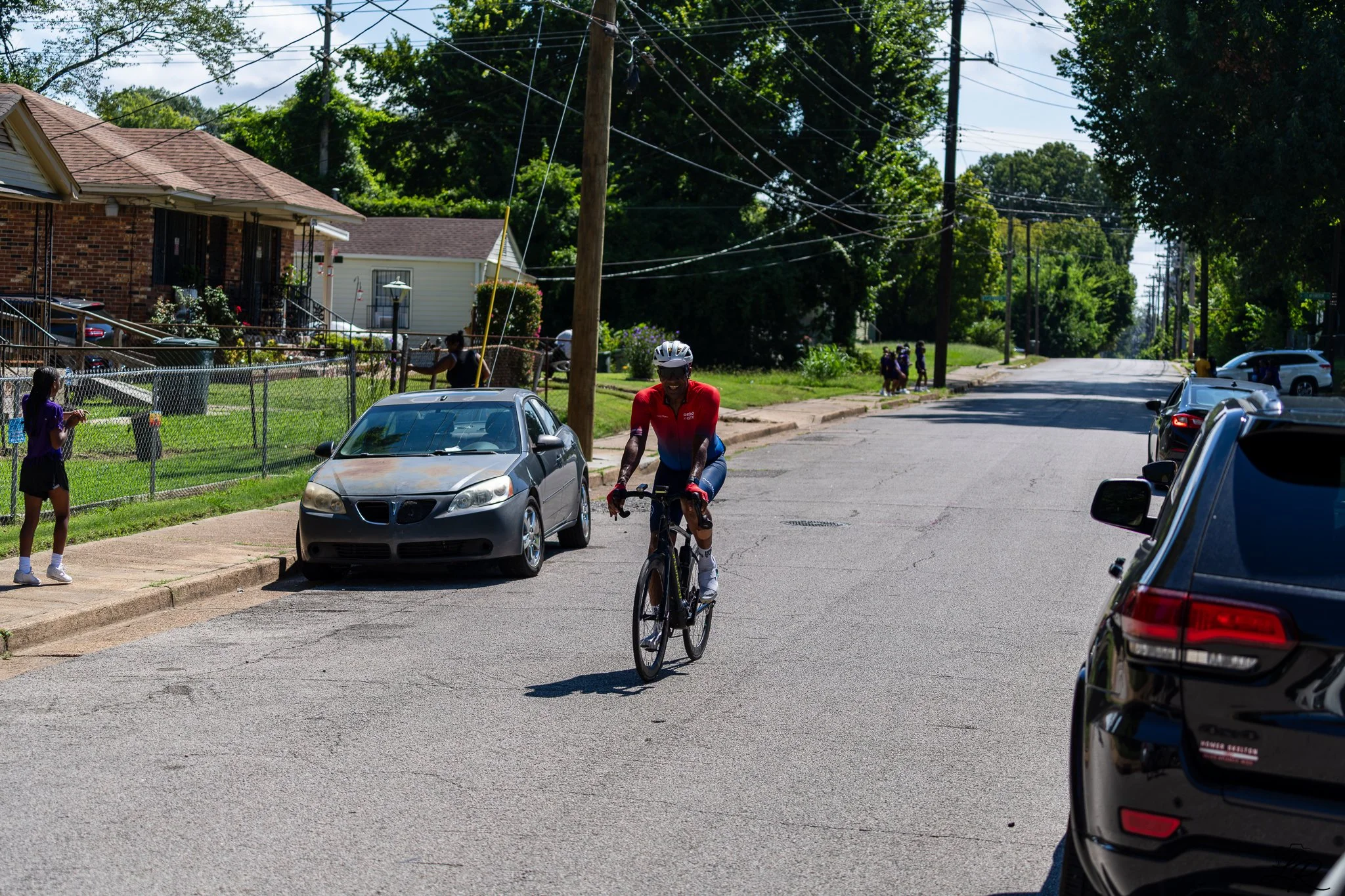 A man riding a bicycle down a residential street with cars parked along the curb, houses in the background, and neighborhood activity visible.