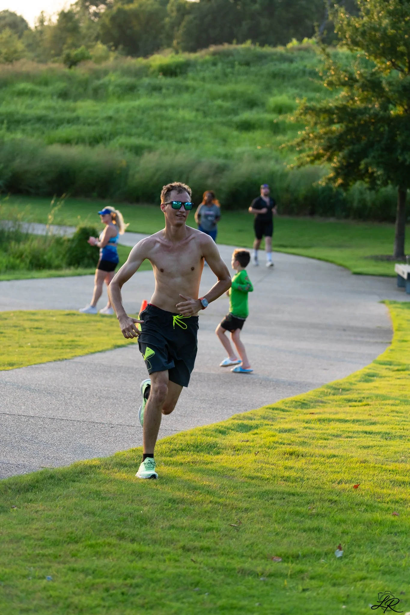 A man running on a park trail with others jogging and walking in the background during sunset. Green grassy area with trees and hill.