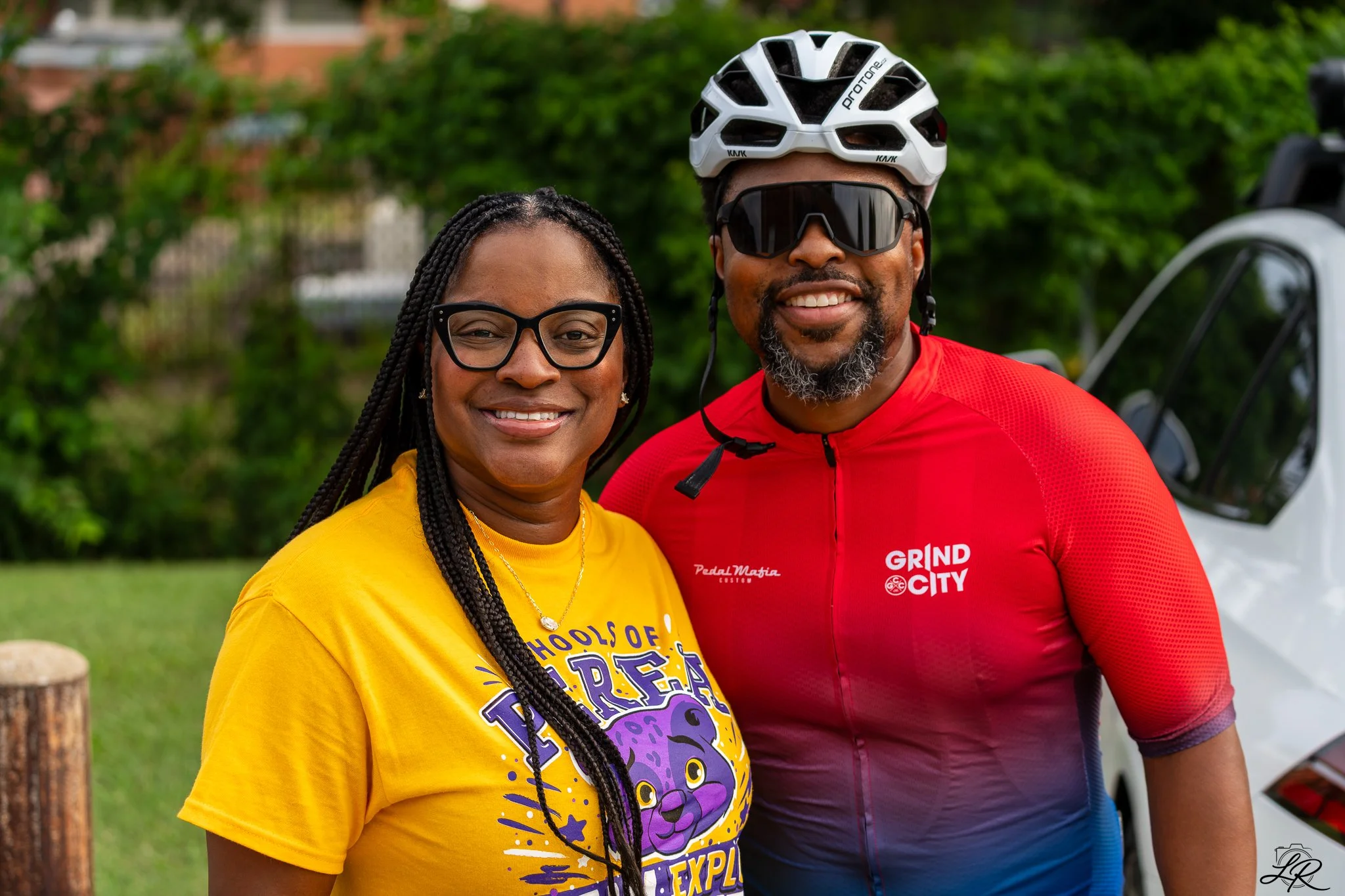 A man wearing a white bicycle helmet, sunglasses, and a red cycling jersey standing next to a woman in yellow shirt with glasses, both smiling outdoors with green foliage in the background.