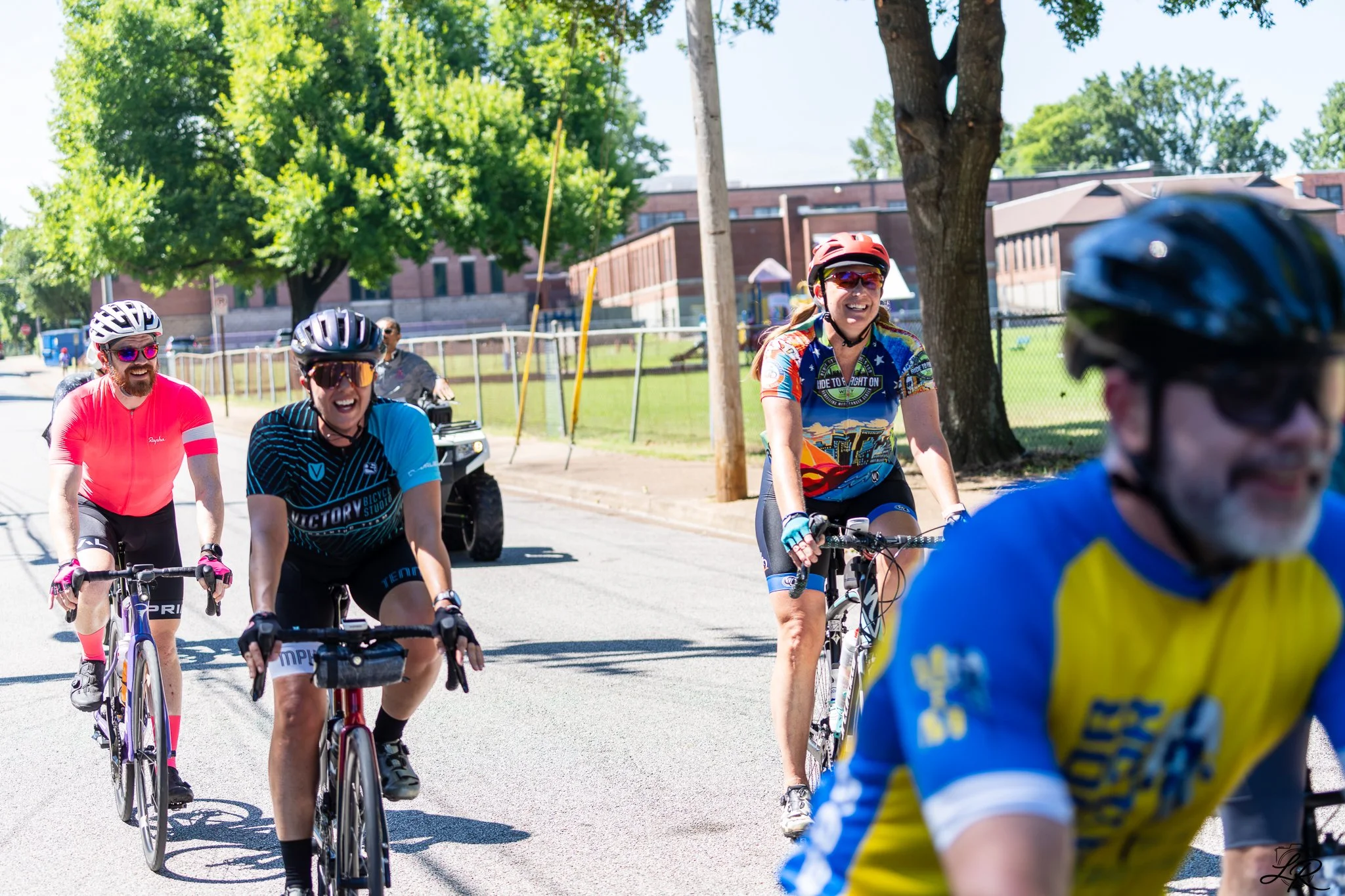 A group of smiling cyclists riding on a sunny street, wearing helmets and colorful cycling uniforms.