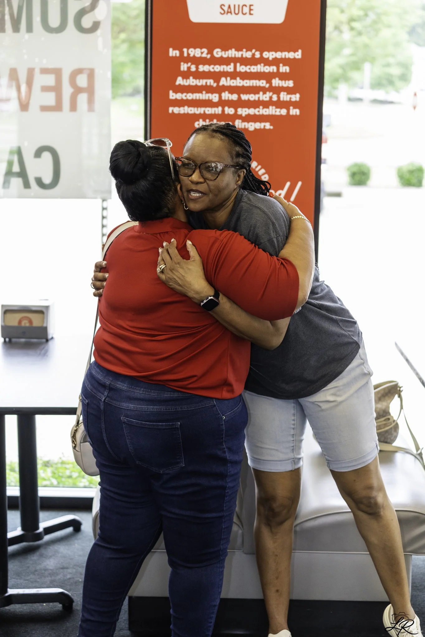 Two woman hugging inside a store near a red sign with text about Guthrie's restaurant history. The woman facing the camera wears glasses, a grey shirt, and light shorts. The woman with her back to the camera wears a red top and jeans.
