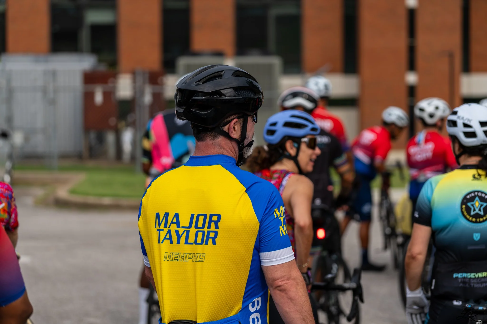 A group of cyclists gathered together, wearing helmets and colorful jerseys, with a focus on a man in a yellow and blue jersey with the name 'Major Taylor' on the back, standing outdoors in front of a brick building.