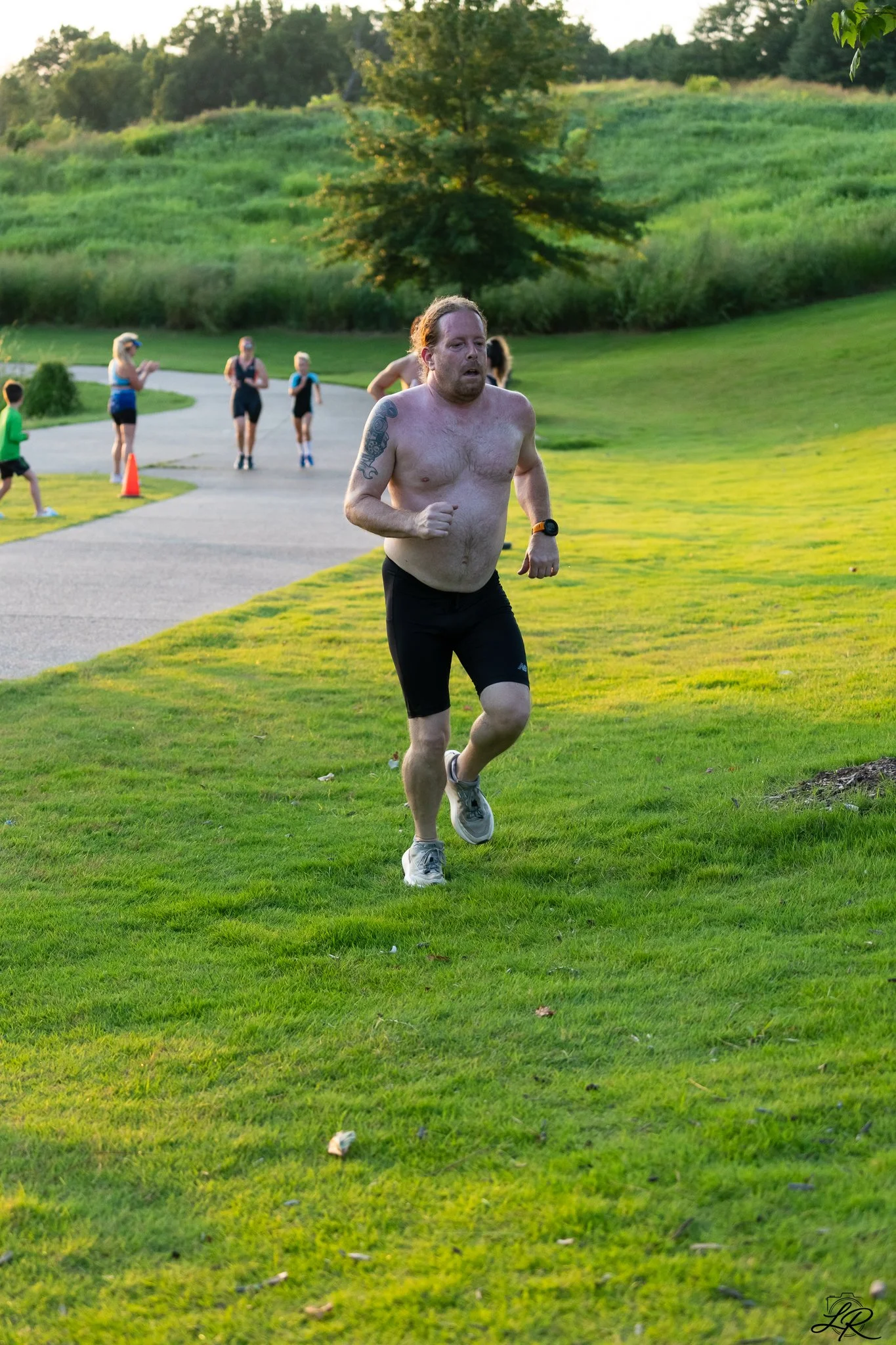 A man with reddish hair running shirtless on a grassy path in a park, with a grassy hill and trees in the background. Several children and a woman are walking or running on the path behind him.