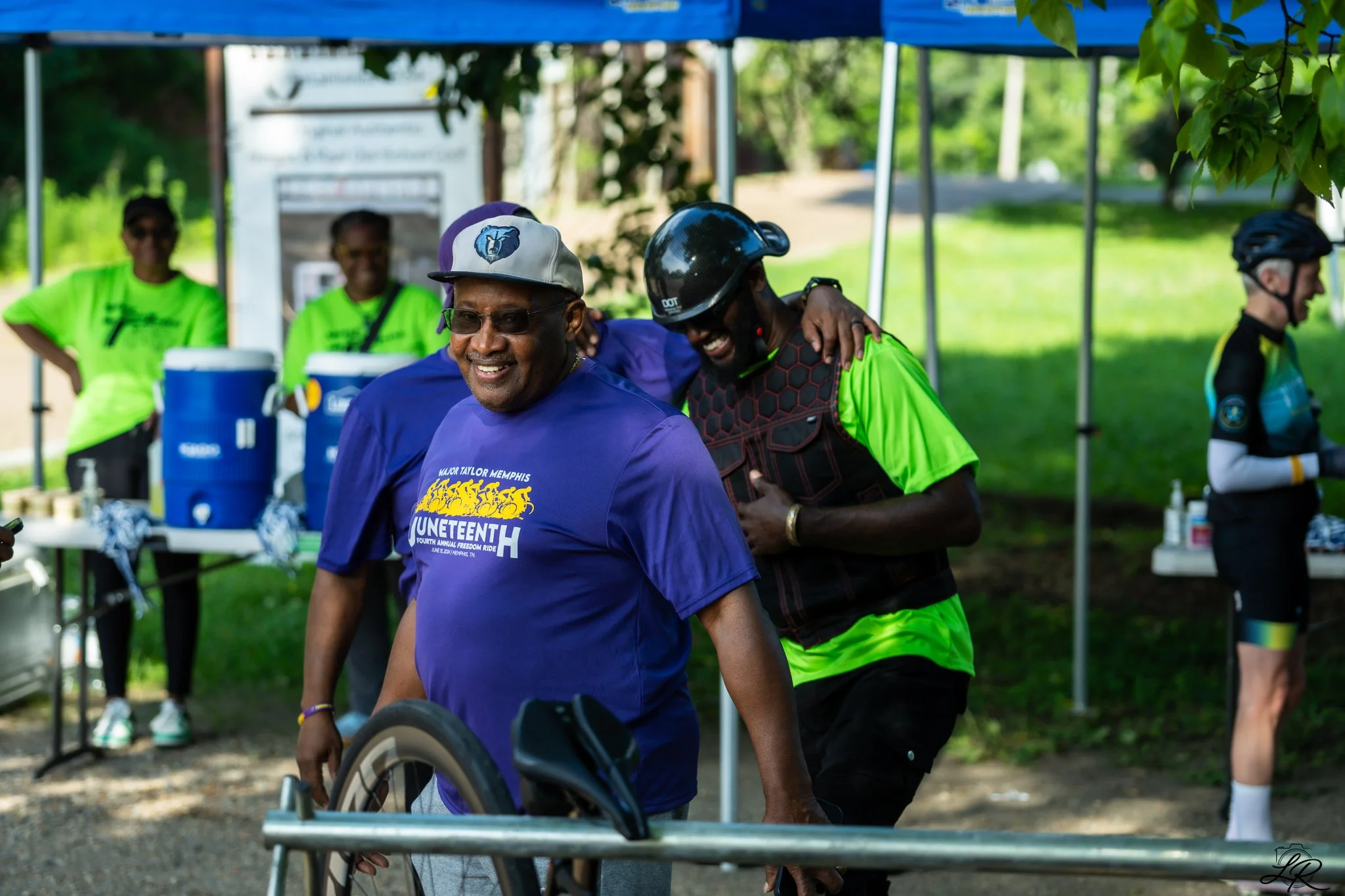 Smiling man in a blue shirt and gray cap being pushed in a wheelchair by another man in a bright green shirt and black helmet at an outdoor event, with people in bright shirts in the background under a blue canopy.