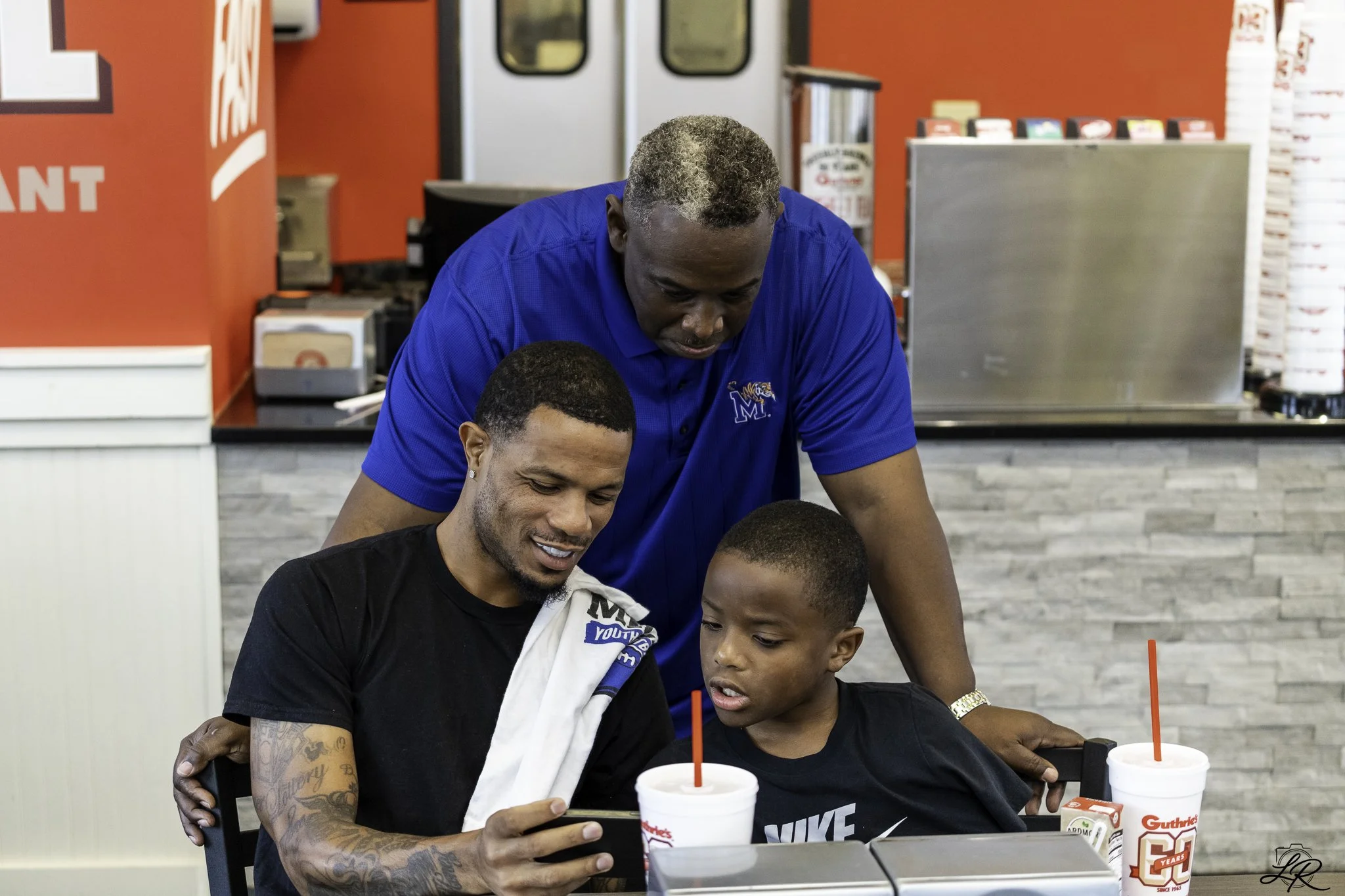 A group of people sitting at a table in a fast food restaurant, looking at a smartphone together, with cups and food items on the table, inside a restaurant with red and gray decor.