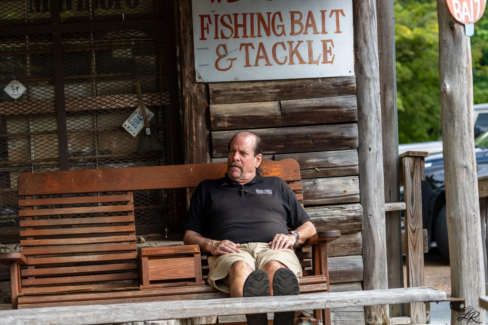 A man with a mustache and beard sitting on a wooden bench outside a rustic building with a sign that reads 'Fishing Bait & Tackle.' He is wearing a black polo shirt and khaki shorts, with his legs stretched out and wearing black shoes. The background