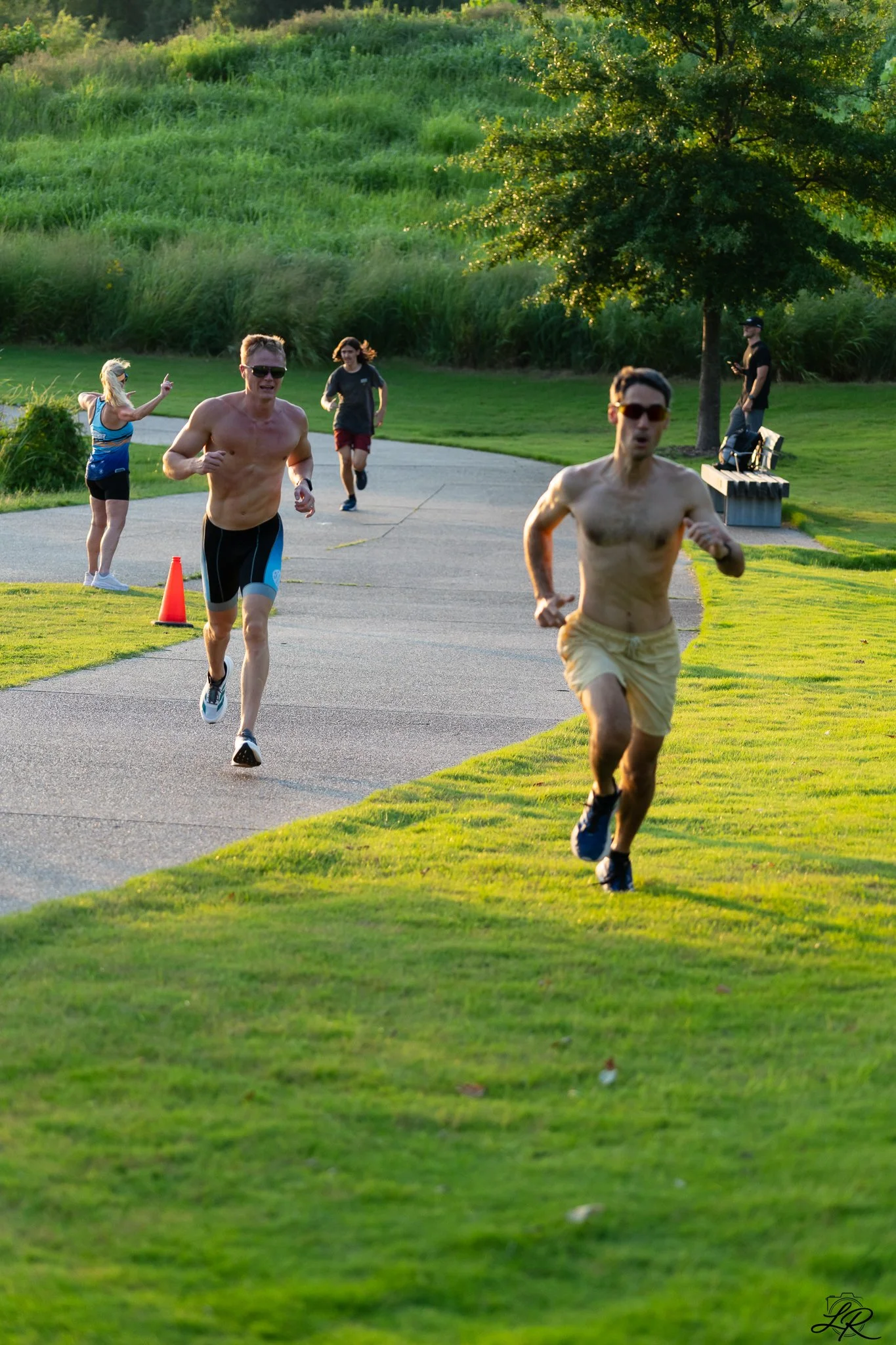 Two shirtless men running on a park path during the evening, with a woman and a man in the background, and a person sitting on a bench under a tree.