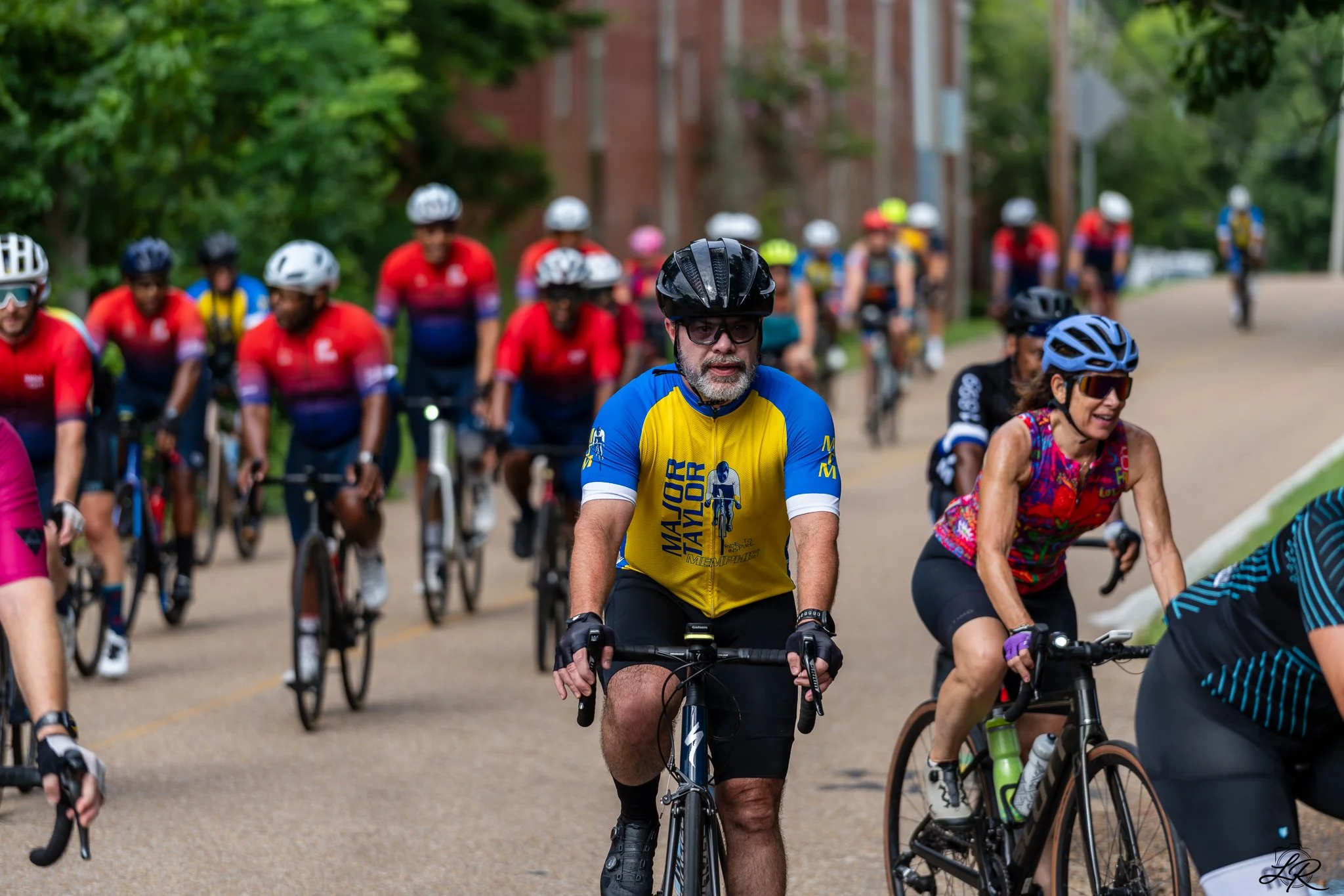 Multiple cyclists riding on a street during daytime, wearing helmets and athletic clothing, surrounded by green trees.