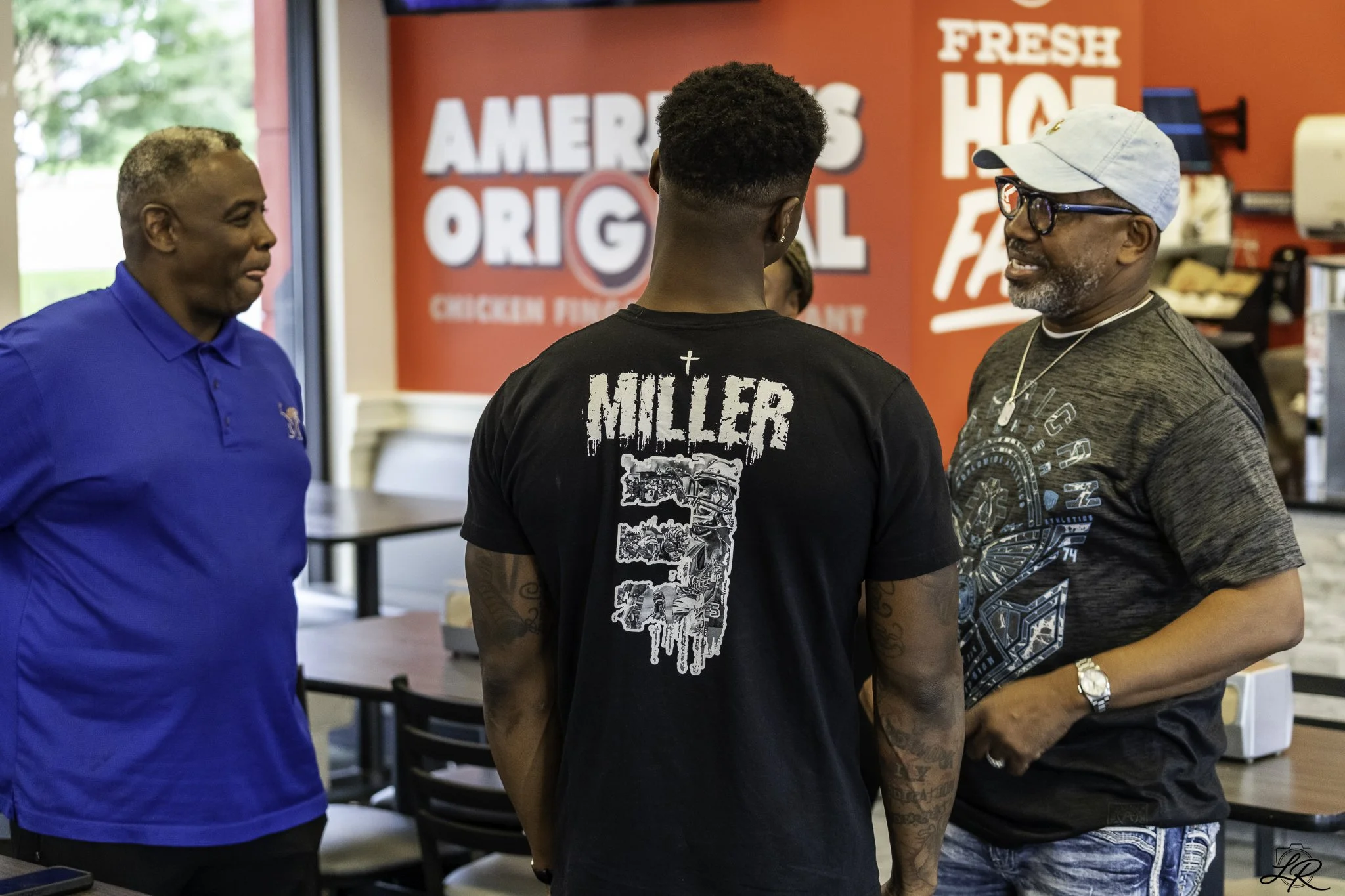 Three men are standing and talking at a fast-food restaurant. The background has a red wall with large white letters that say 'AMERICA'S ORIGINAL' and 'FRESH HOT'. One man is wearing a blue shirt, the second man has a black shirt with 'Miller' writte