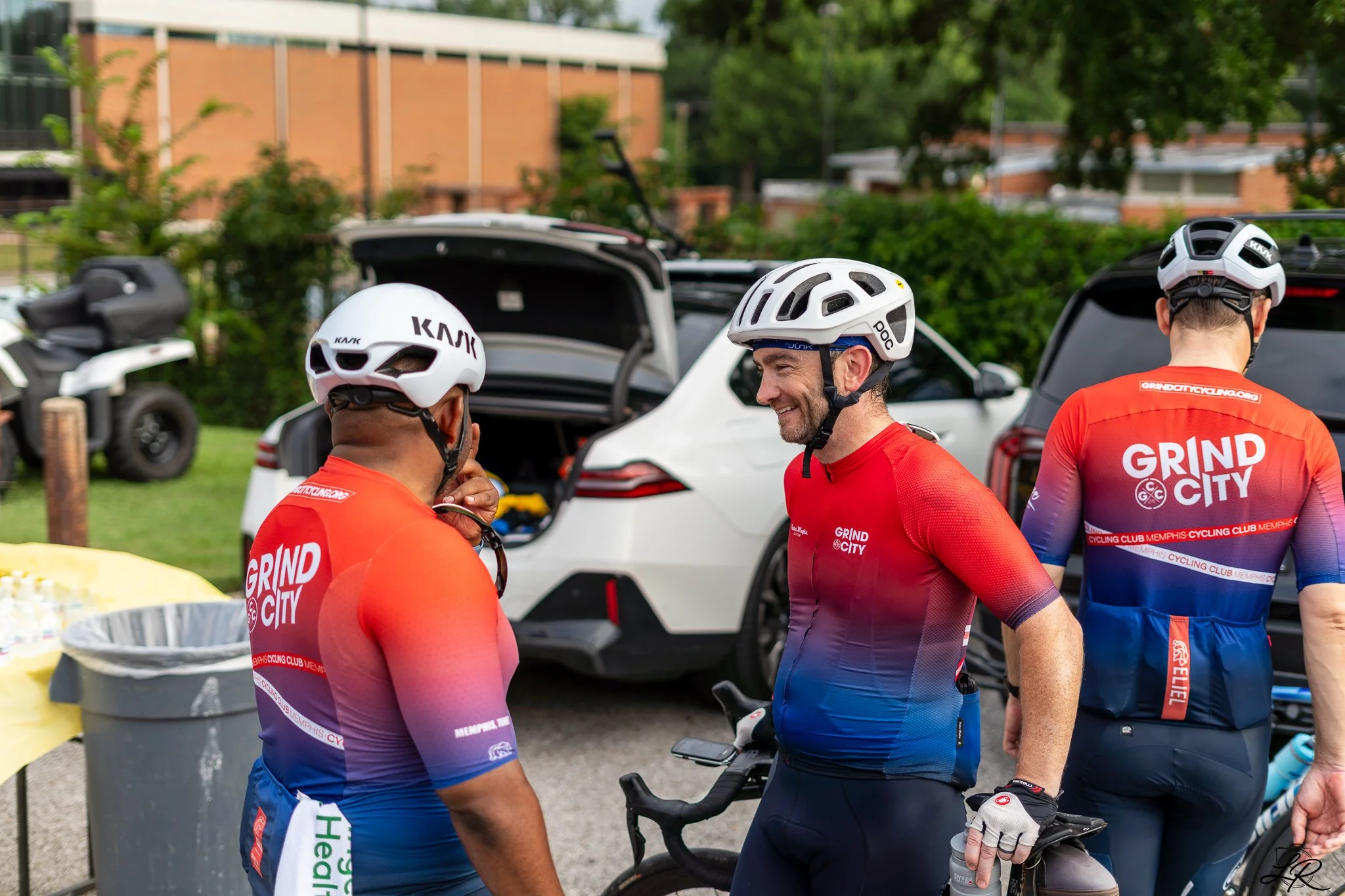 Cyclists in red and blue jerseys talking and preparing to ride bikes, parked cars, and lush green trees in the background.