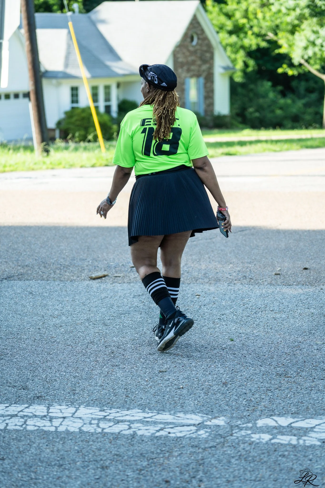 A person with dreadlocks, wearing a black cap, bright green sports shirt with black writing and a number 13, a black skirt, black and white striped knee-high socks, and black sneakers, walking on a cracked street.