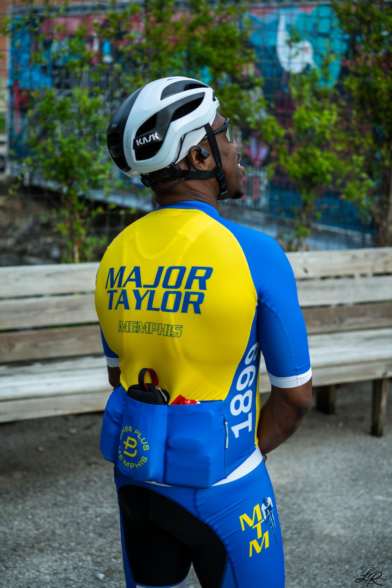 Side view of a male cyclist wearing a white helmet, sunglasses, a yellow and blue Memphis Major Taylor cycling jersey, and blue cycling shorts, standing outdoors near a wooden fence with trees and colorful graffiti in the background.
