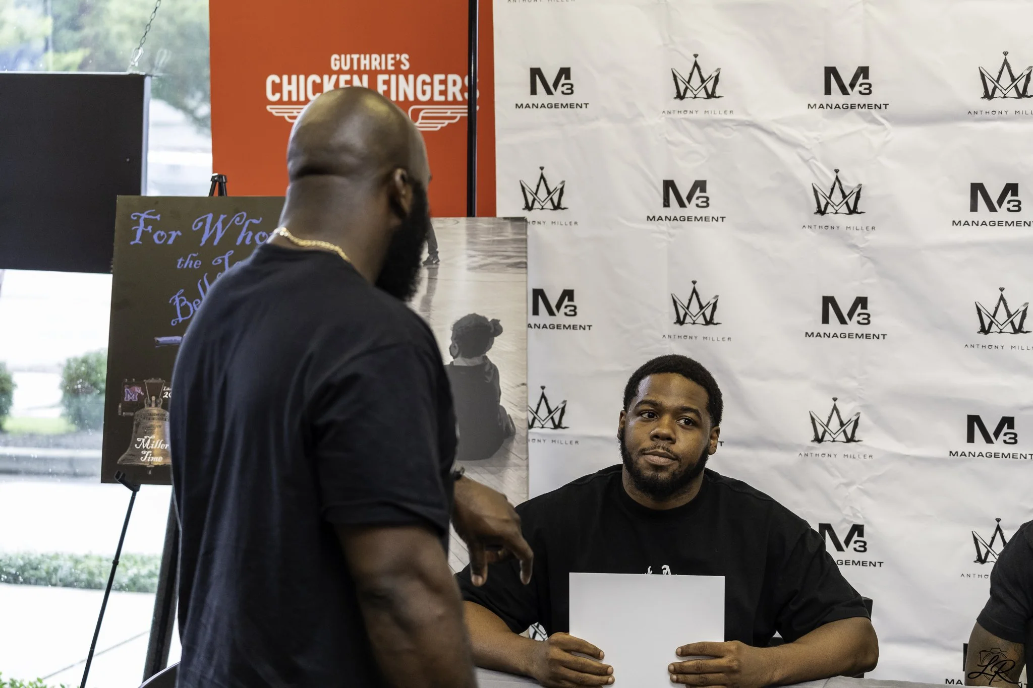 Two men at a signing event with a backdrop featuring logos of 'M3 Management' and 'Anthony Miller.' One man, with a shaved head, is standing and facing away, while the other man, with short hair and a beard, is seated, holding a pen and looking at th