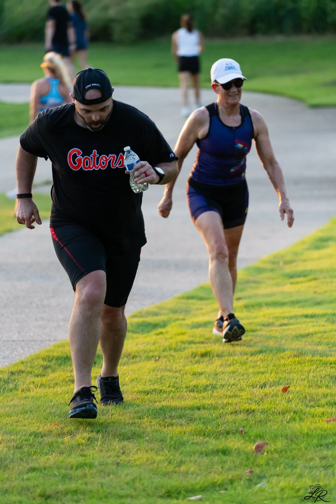 A group of people, including a man in a black T-shirt with 'Gators' written on it and a woman in a blue sports tank top, are walking or jogging on a grassy path in a park or outdoor area during the late afternoon or early evening. Other individuals c