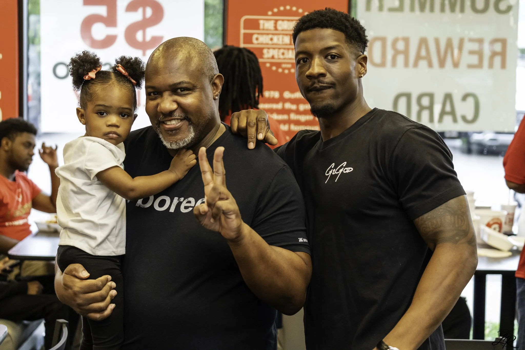 A young man making a peace sign and a man holding a young girl in his arms inside a restaurant. The girl is looking at the camera with a serious expression and has her hair styled in two puffs with pink bows. The background features red and white sig