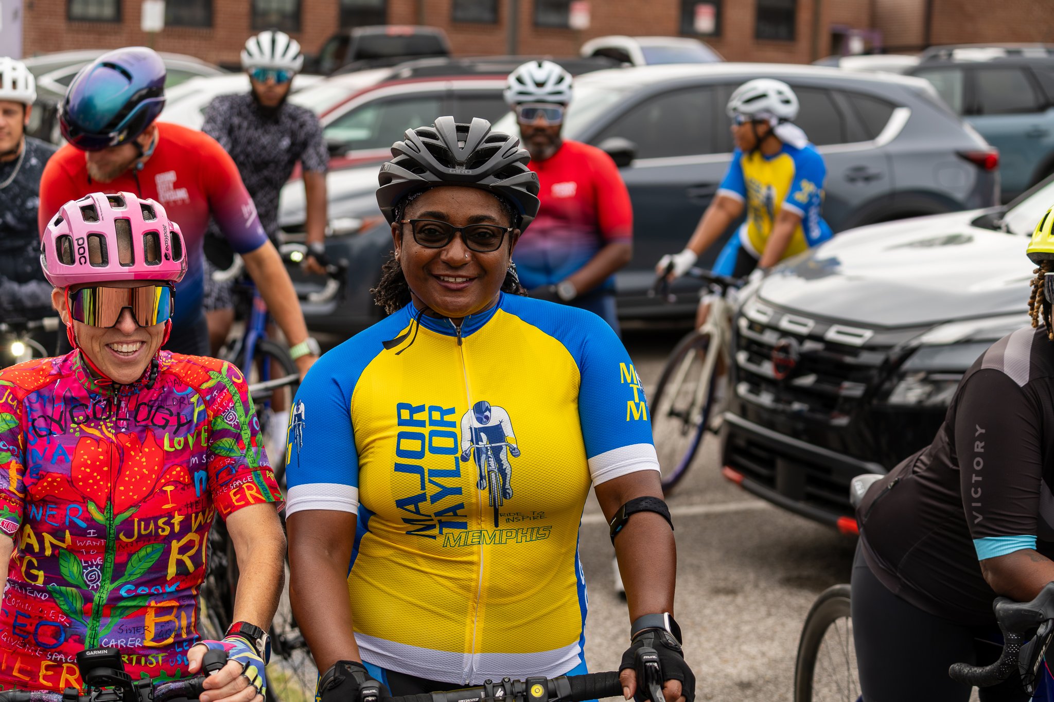 Group of cyclists in colorful jerseys and helmets standing in a parking lot next to cars.