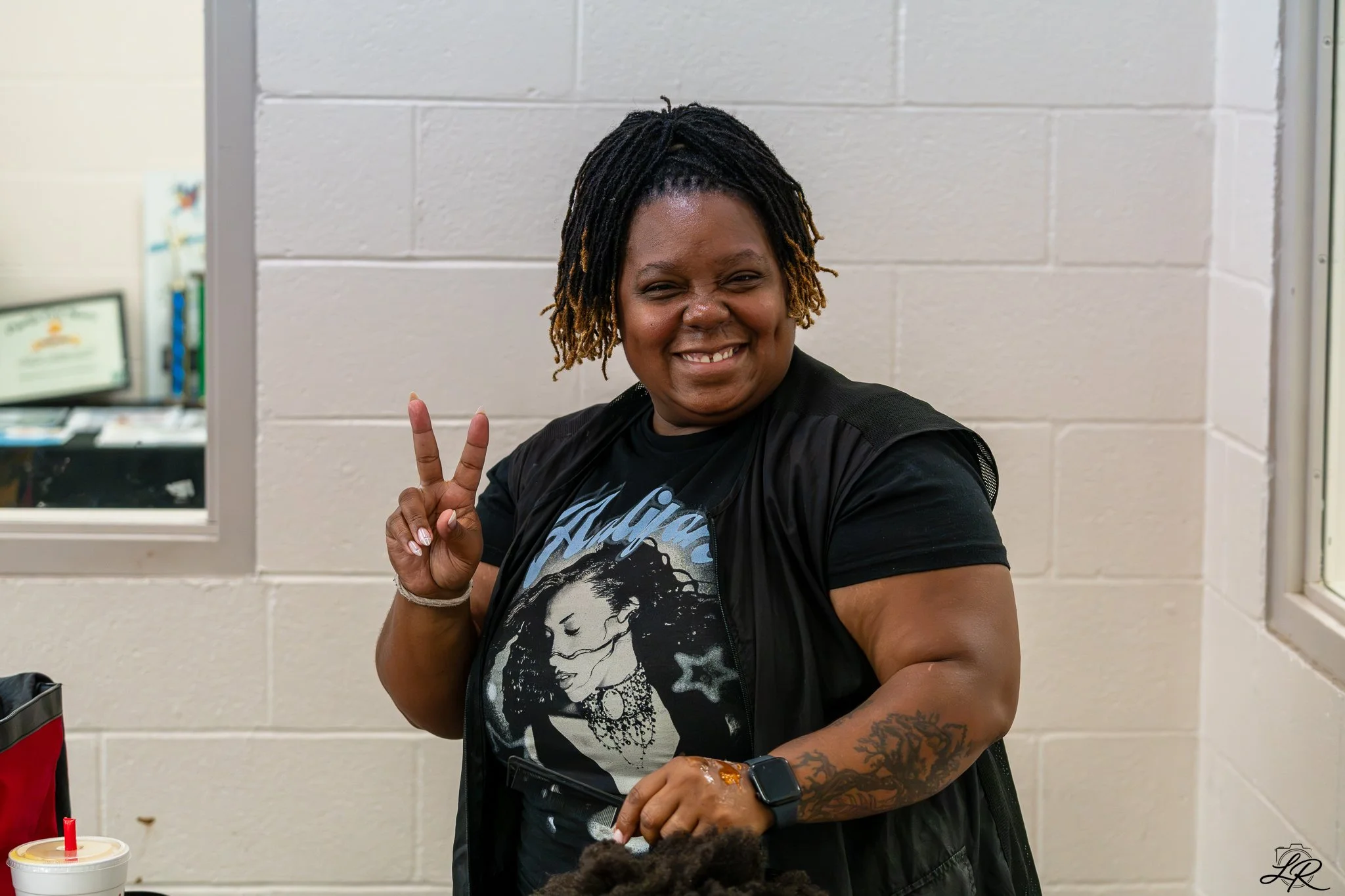 A woman with dreadlocks and tattoos is smiling and making a peace sign with her fingers. She is wearing a black T-shirt with a woman's face printed on it and a black jacket. There is a drink with a red straw on the table in front of her. The backgrou