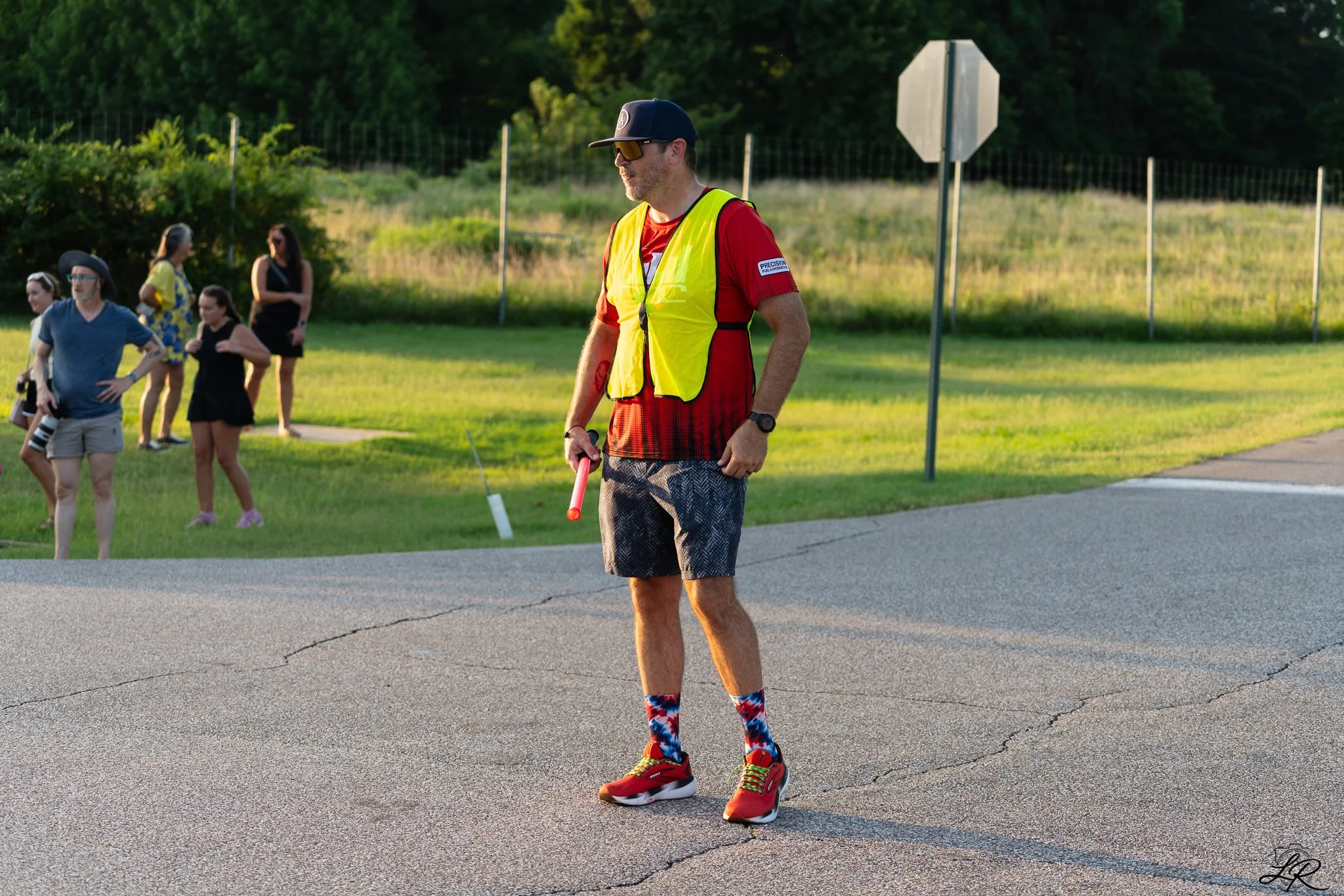 A man walking on the roadside wearing a bright yellow safety vest, red shirt, patterned shorts, red running shoes, colorful socks, a black cap, and sunglasses, holding a red walking stick, with a group of people in the background near a grassy area w