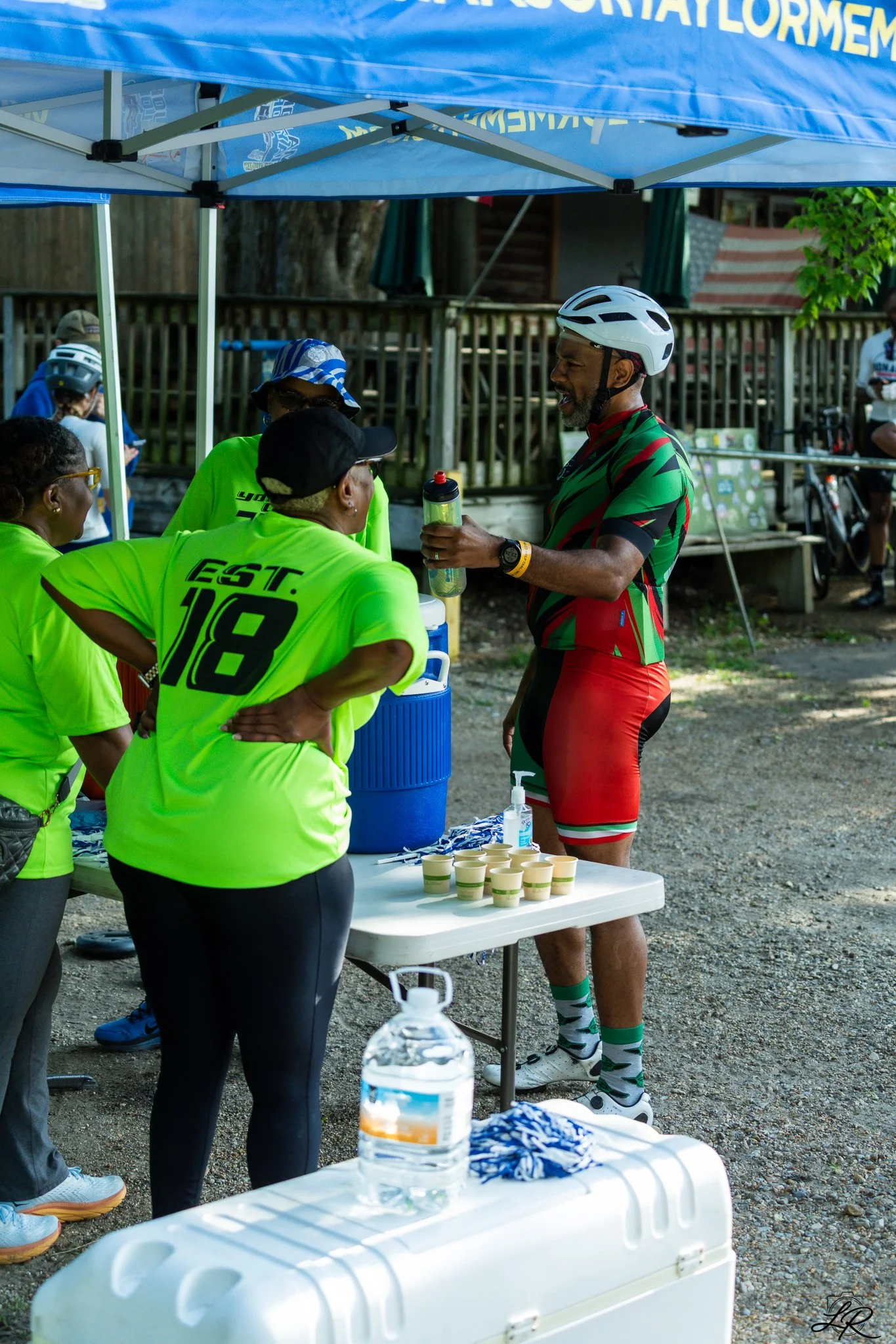 A group of people at a race event, with the main focus on a man in a cycling outfit wearing a helmet and holding a water bottle, talking to a woman in a bright yellow shirt. They are under a blue canopy, and there are cups, water bottles, and other s
