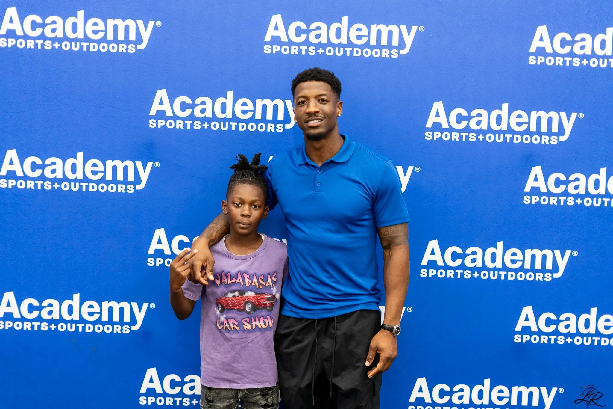 A man and a young girl standing in front of a blue backdrop with white text that reads 'Academy Sports + Outdoors'. The man is wearing a blue polo shirt and has tattoos on his right arm, while the girl is wearing a purple t-shirt with a graphic of a 